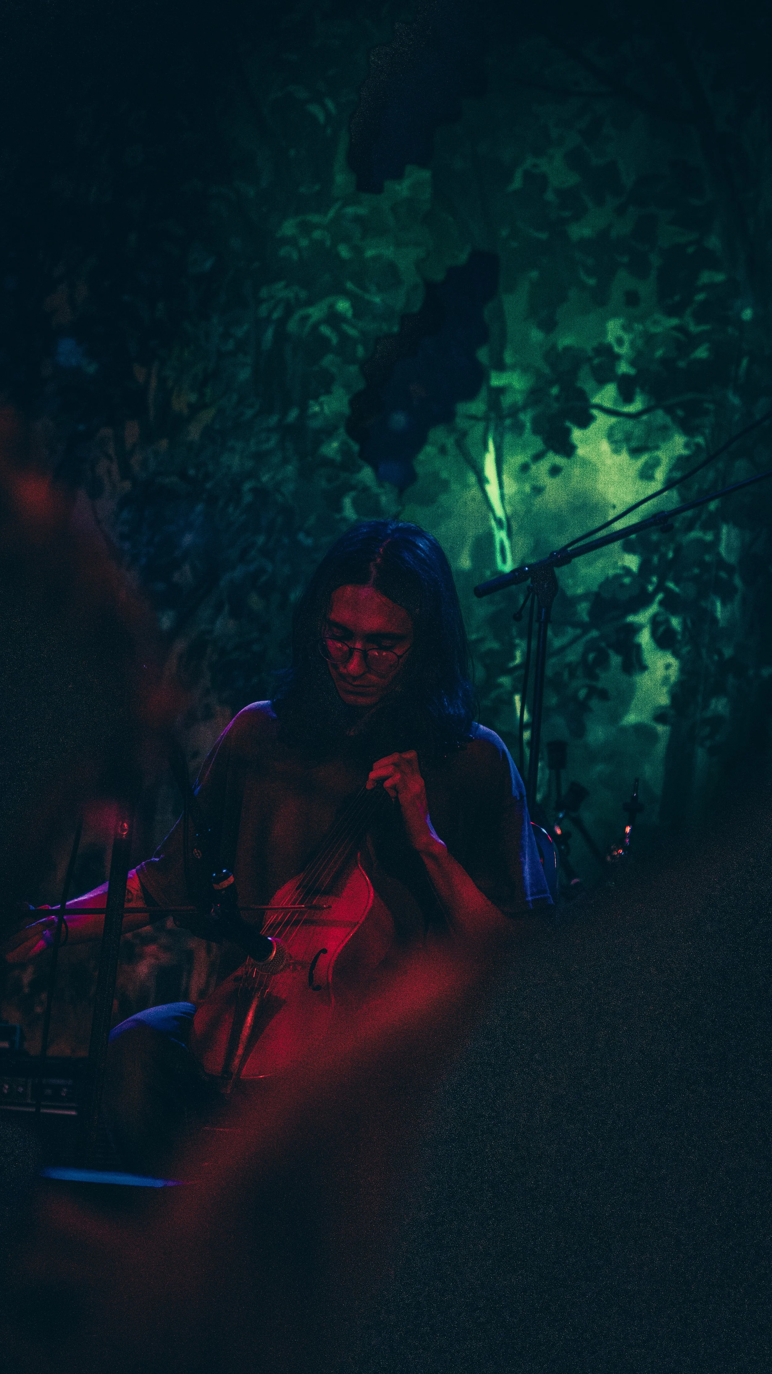 A woman sits in a dimly lit room, playing a string instrument, with colorful lights and abstract patterns projected on the wall behind her.
