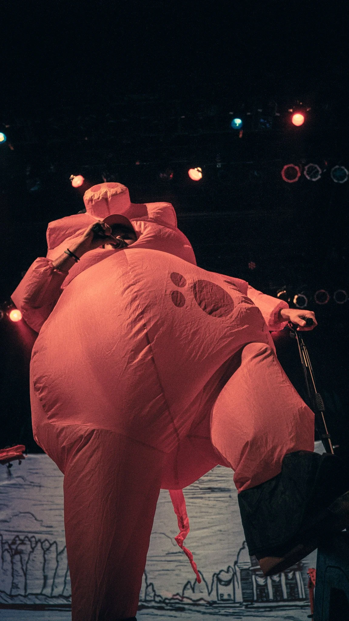 Performer in a pink inflatable costume resembling a pig, holding a microphone on stage with colorful lights and a cityscape backdrop.