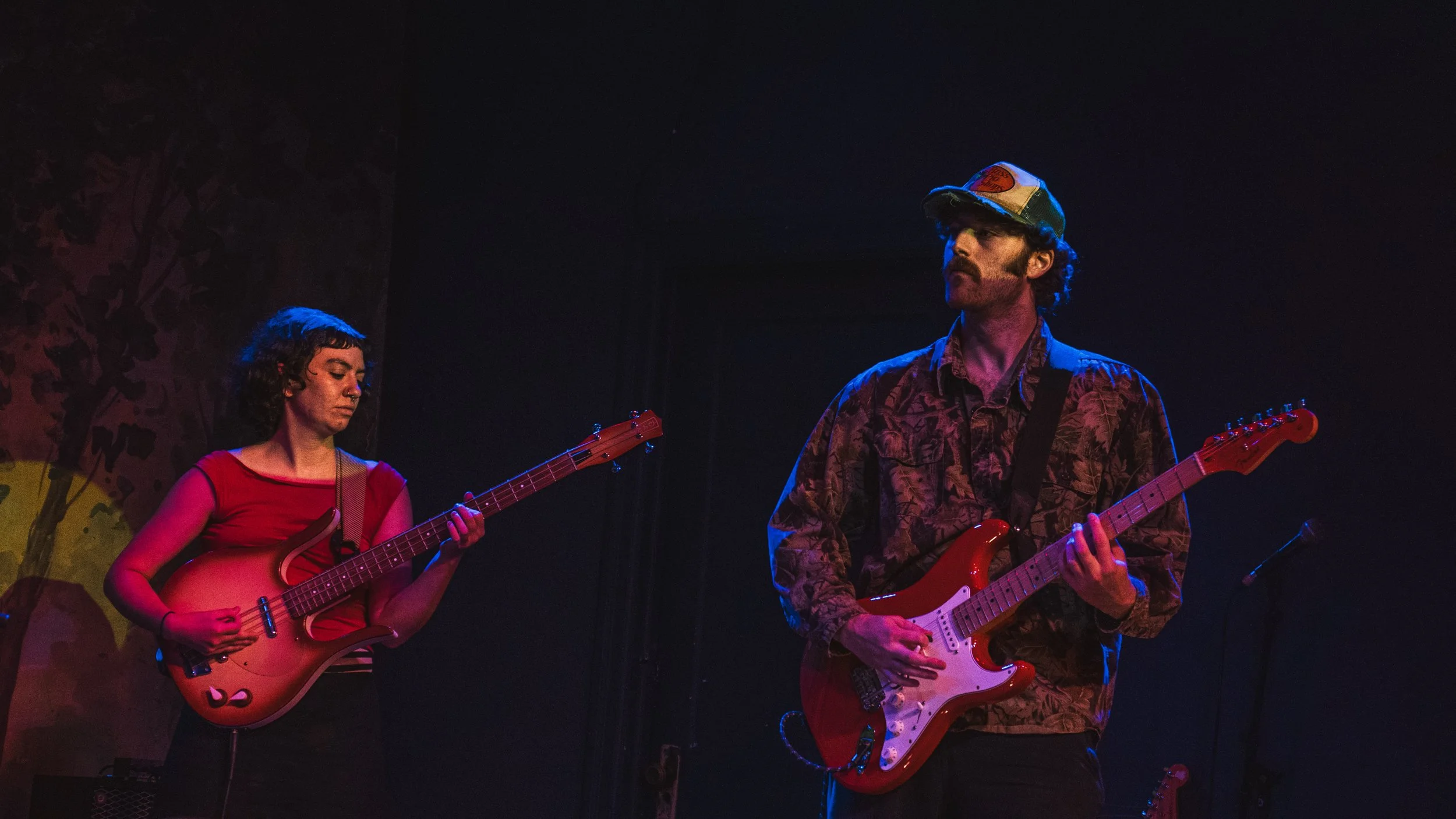 Two musicians performing on stage with guitars, a woman on the left and a man on the right, under colorful stage lighting.