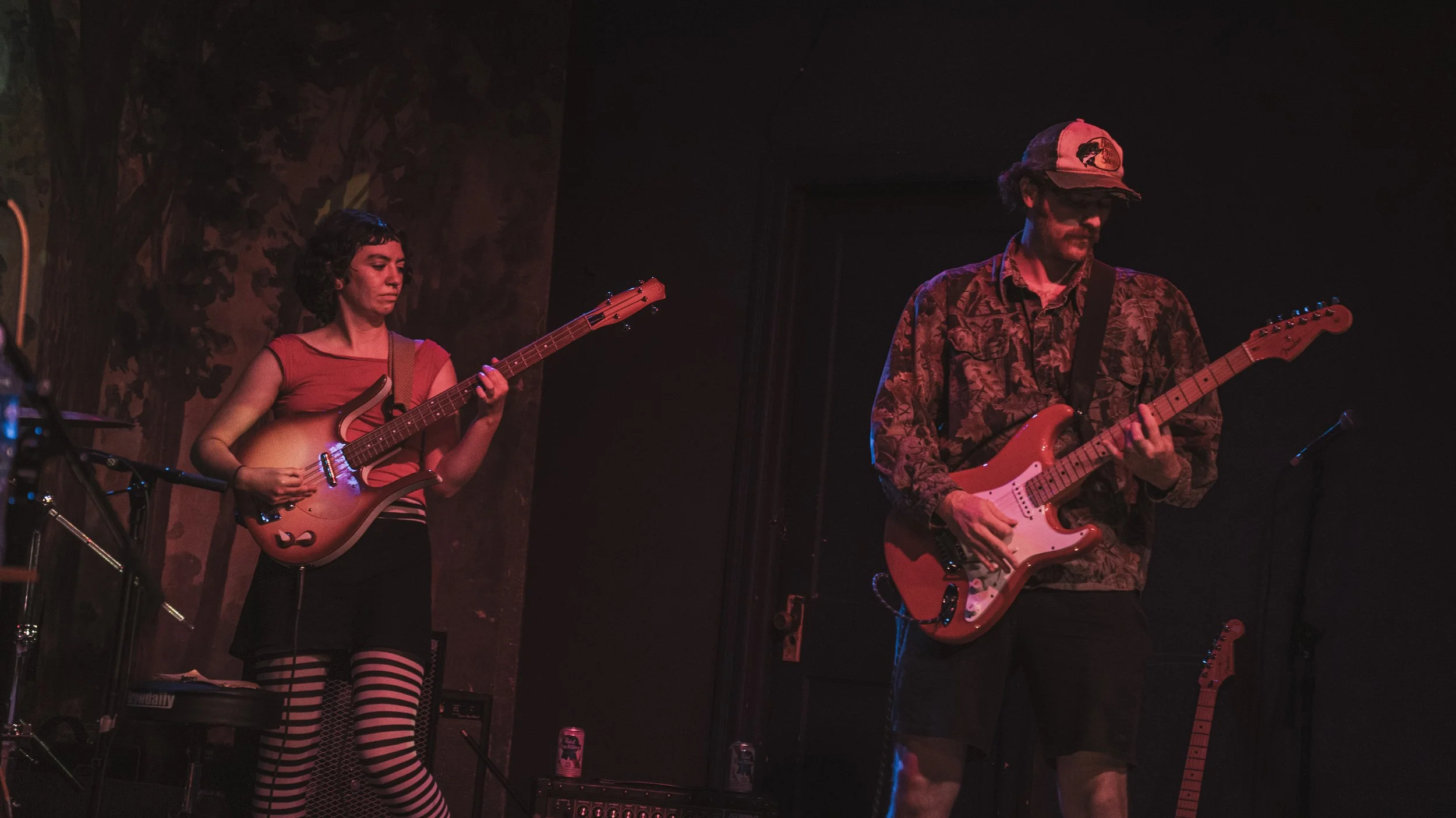 Two musicians performing on stage: a woman playing a sunburst electric bass guitar, wearing a red top and striped leggings; a man playing a red electric guitar, wearing a patterned shirt, a cap, and glasses, with a dark background.
