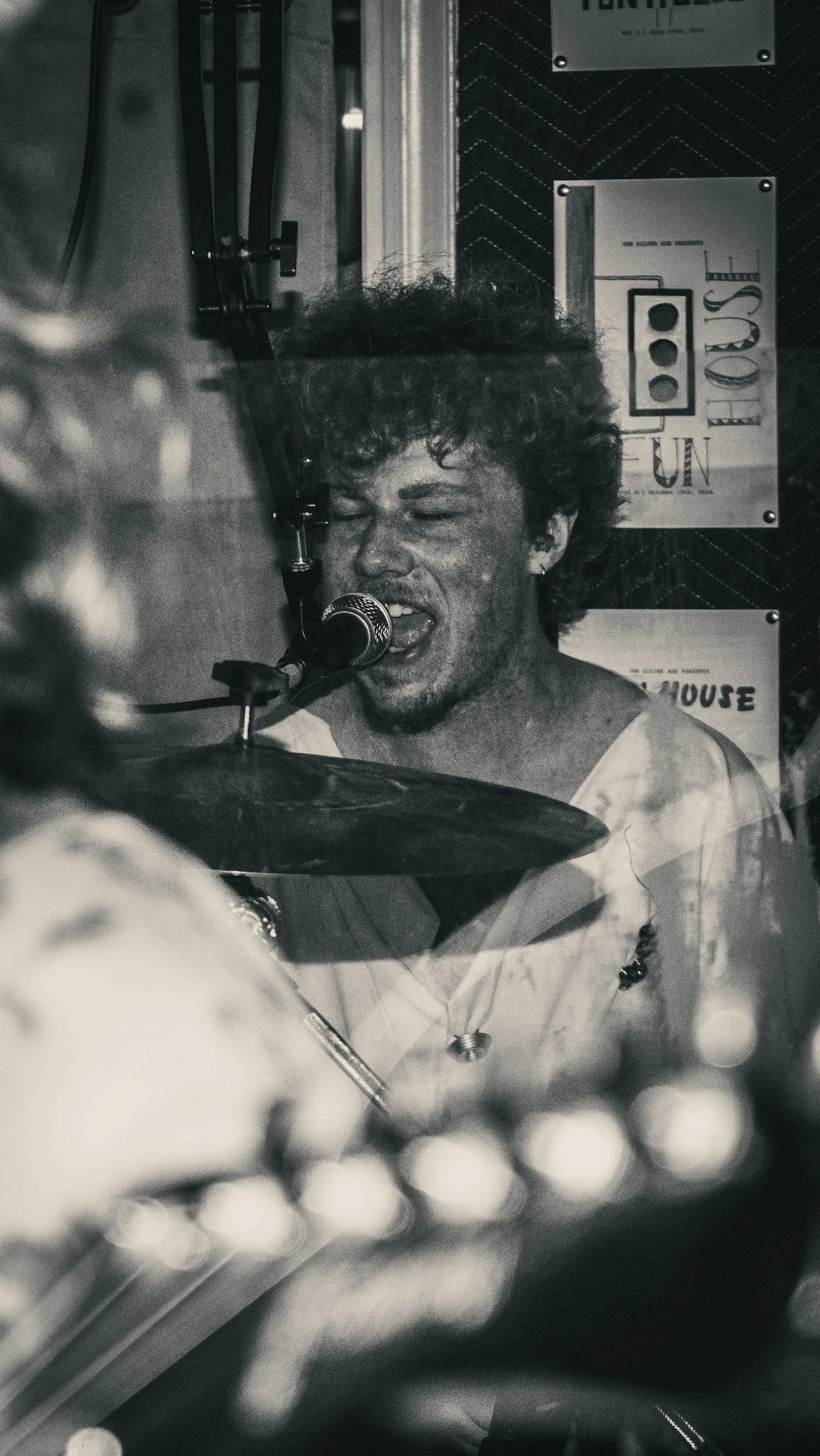 A person singing into a microphone while playing drums, with posters on the wall behind them.