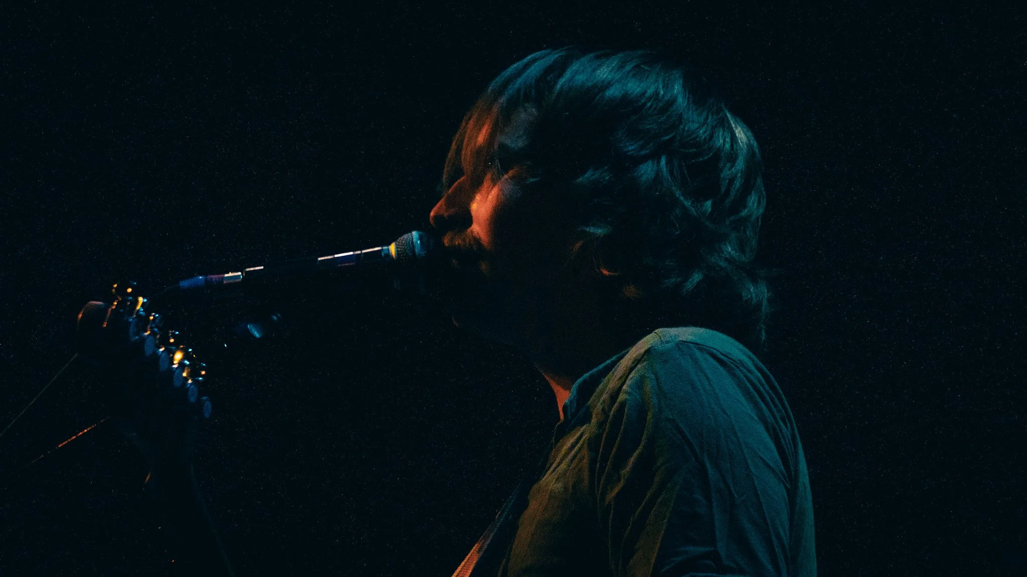 A person with long hair playing an electric guitar in a dark setting, illuminated by subtle blue and orange lighting.