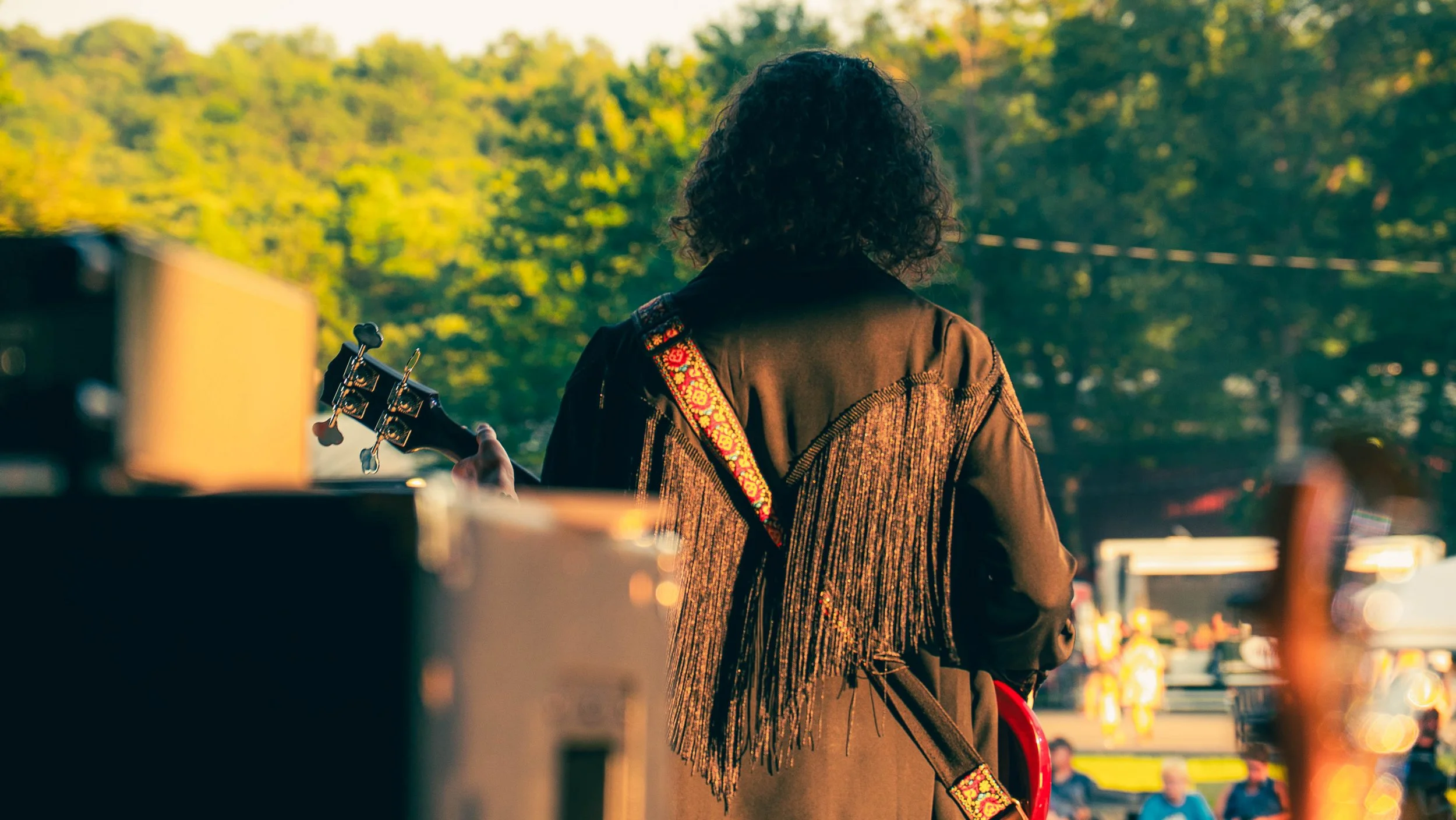 Person with curly hair playing guitar outdoors on a sunny day surrounded by trees and traffic.