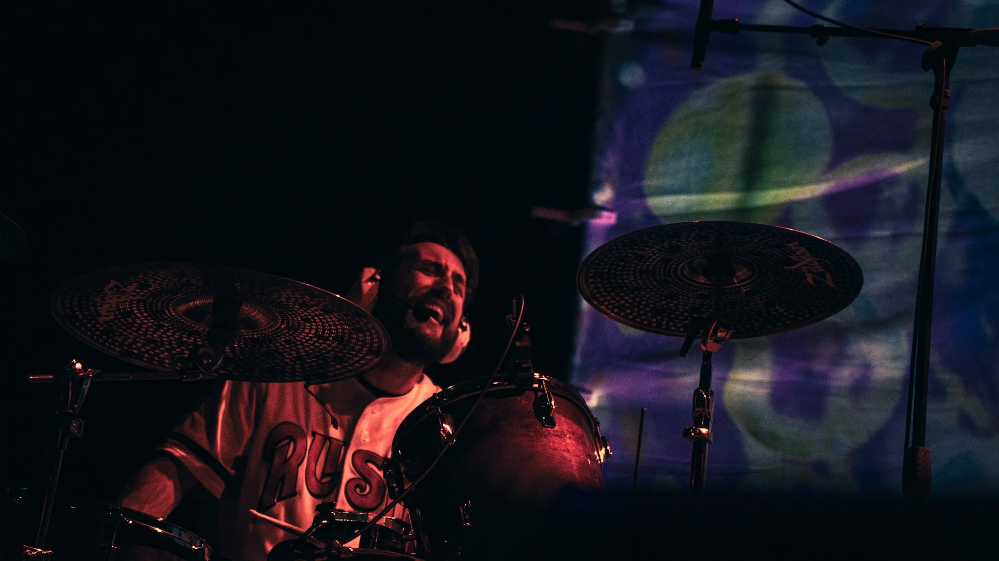 A person playing drums, smiling, with colorful lighting and sound equipment in a dark setting.