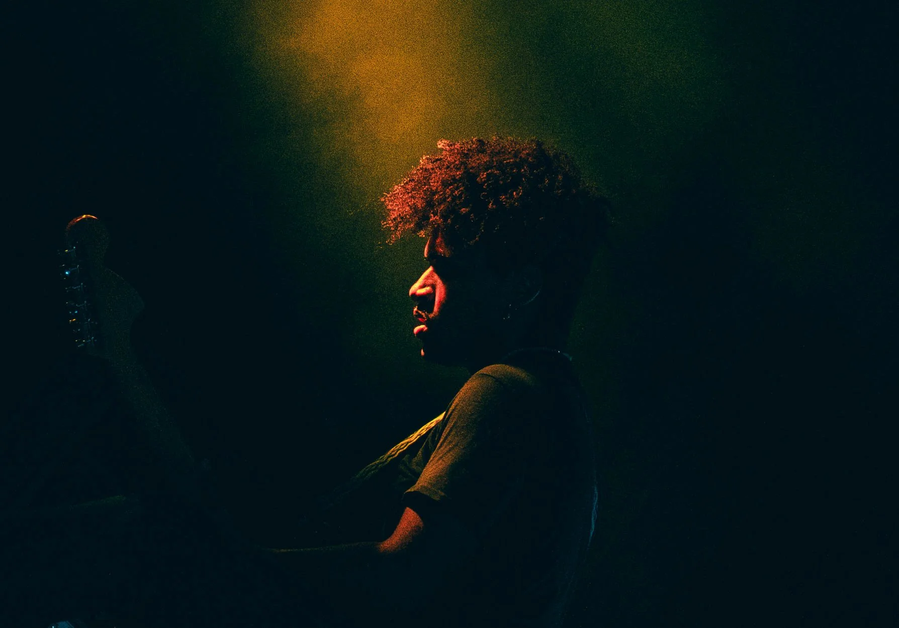 A person with curly hair holding a microphone on stage, illuminated by colorful stage lighting in a dark setting.