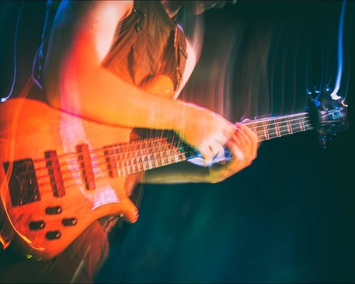 A person playing an orange electric bass guitar on stage with colorful stage lighting.