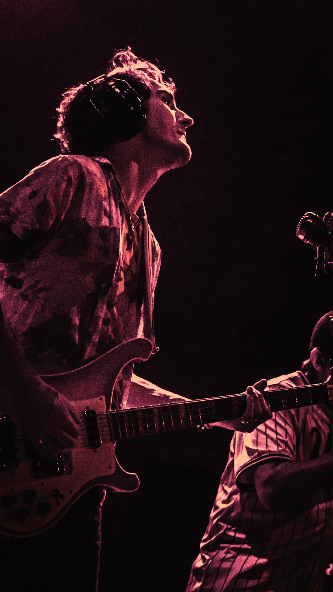 Musician with curly hair wearing headphones, playing an electric guitar on stage, with dark background and purple lighting.