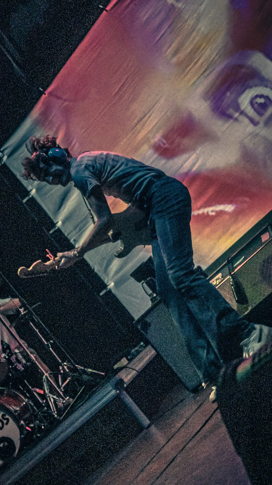 A woman wearing headphones and a short-sleeved shirt, standing on stage with a microphone in one hand. The background features a colorful, illuminated backdrop with a stylized face or logo. The setting appears to be a performance or concert.