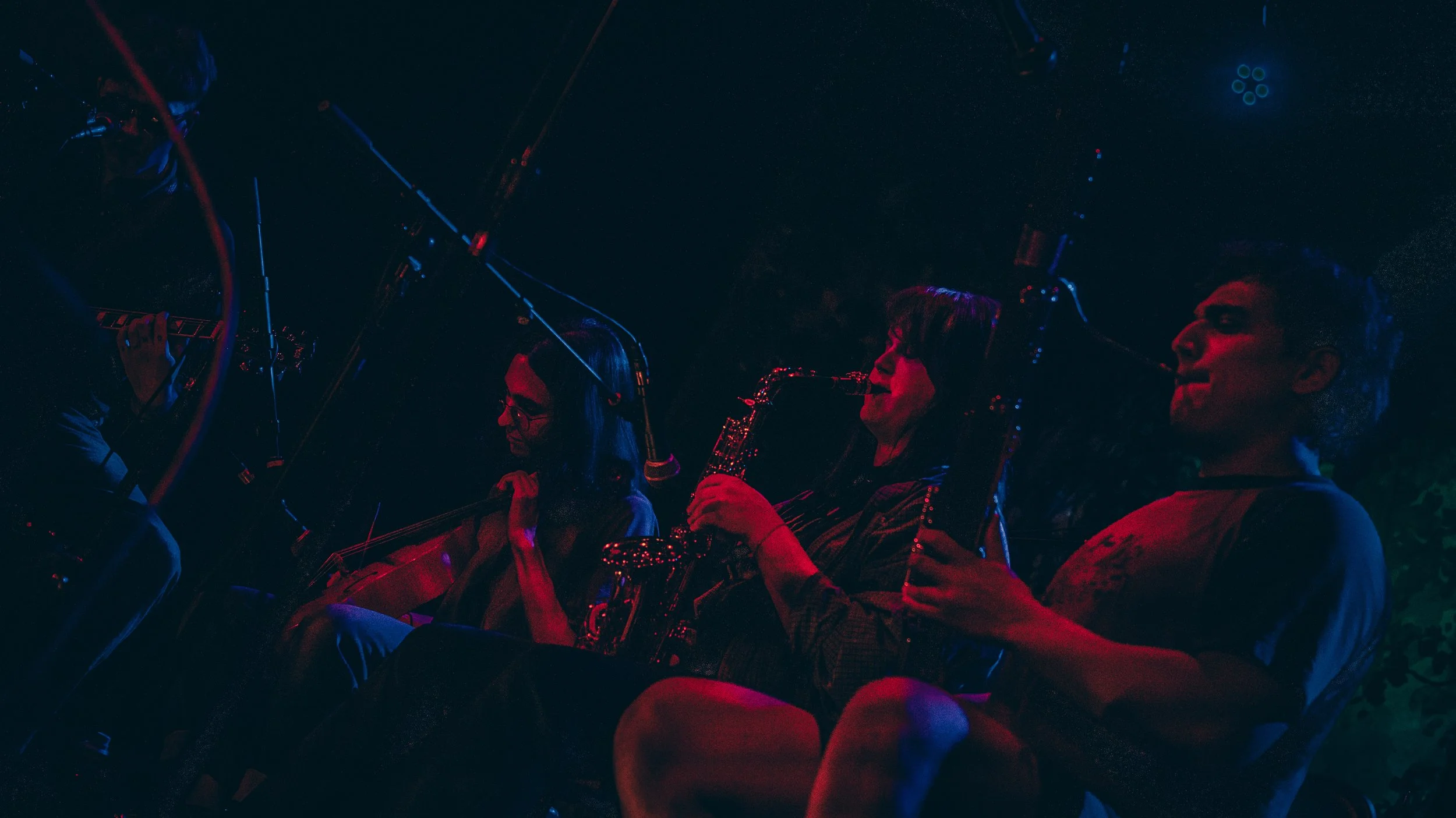 Four musicians playing instruments in a dark, illuminated setting with colorful lighting.