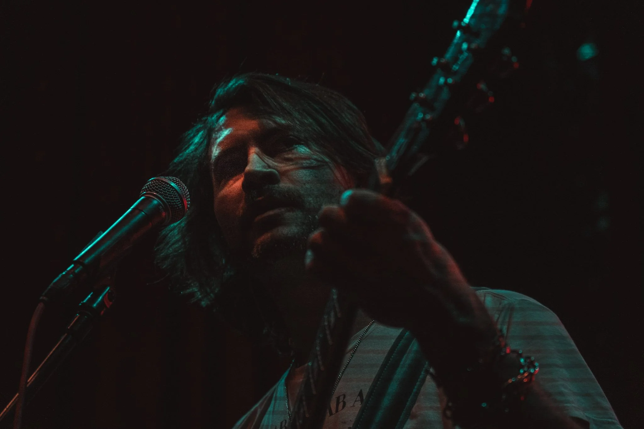 A male singer performing on stage with a microphone in front of him, dimly lit with red and blue lighting, and a guitar visible in the background.