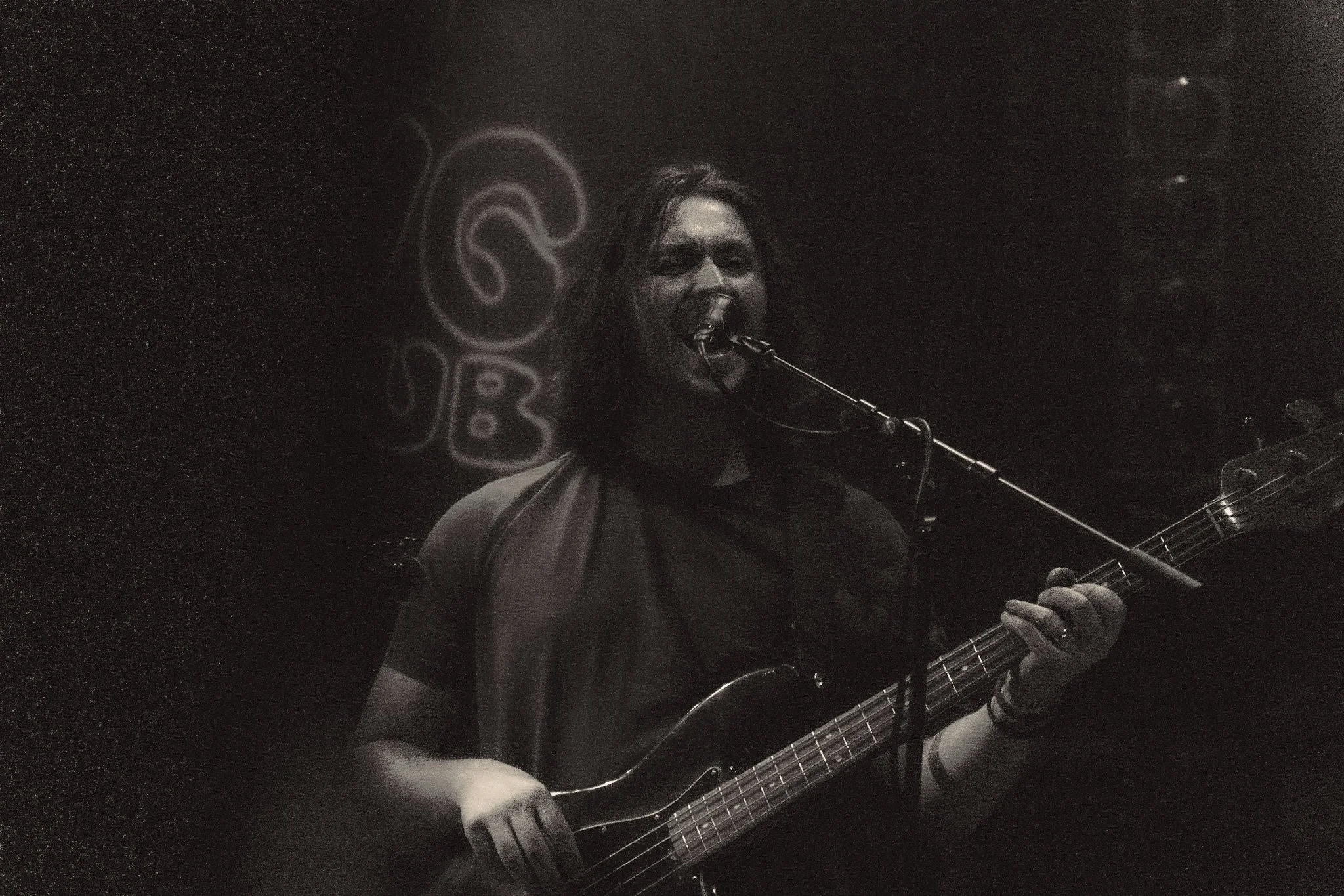 A person with long hair and a mask over their face is playing an electric guitar and singing into a microphone in a dark setting. There is a neon sign or art piece in the background.