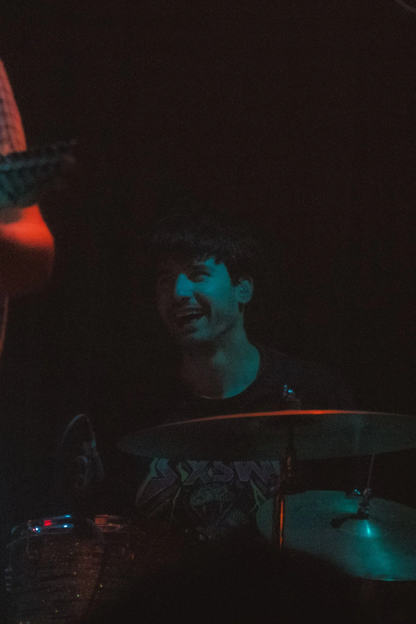 A man with curly hair illuminated by blue light, smiling in a dark environment, likely DJing or performing at a nightclub or party.