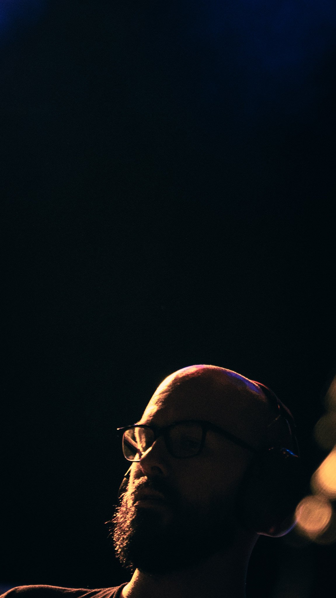A person with glasses and a beard, illuminated by a soft light, appears in a dark background.