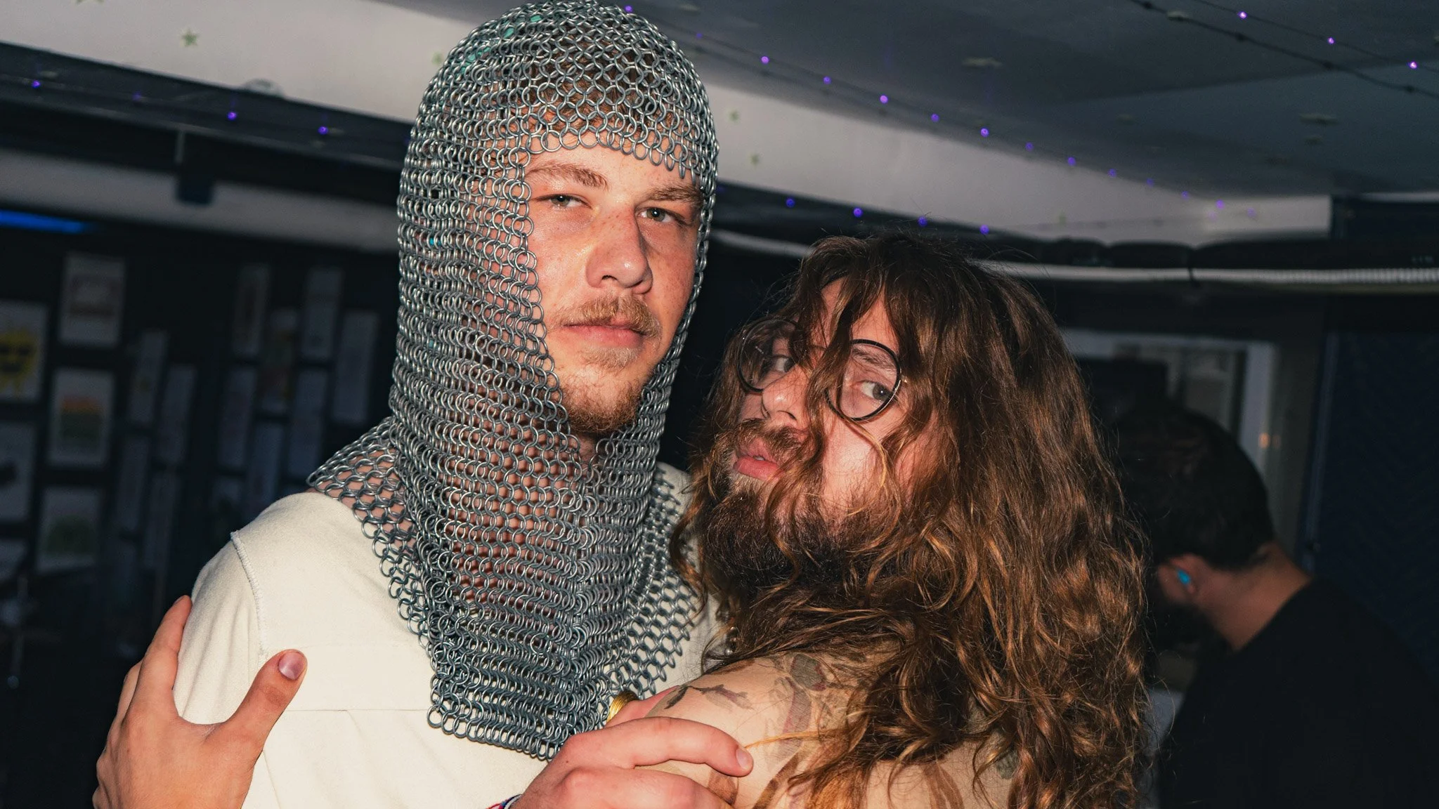A man wearing chainmail armor on his head and shoulders, standing next to a woman with long, curly hair and glasses, in an indoor setting with dim lighting and purple decorative lights on the ceiling.