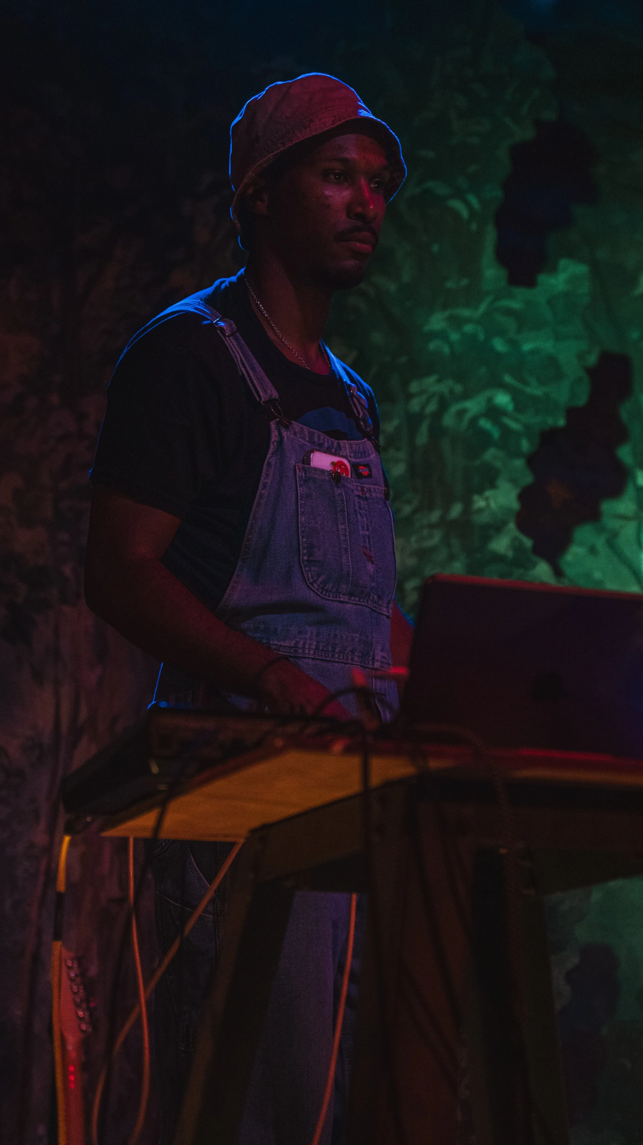 A young man wearing a bucket hat and overalls stands in a dimly lit environment with multicolored neon lights.