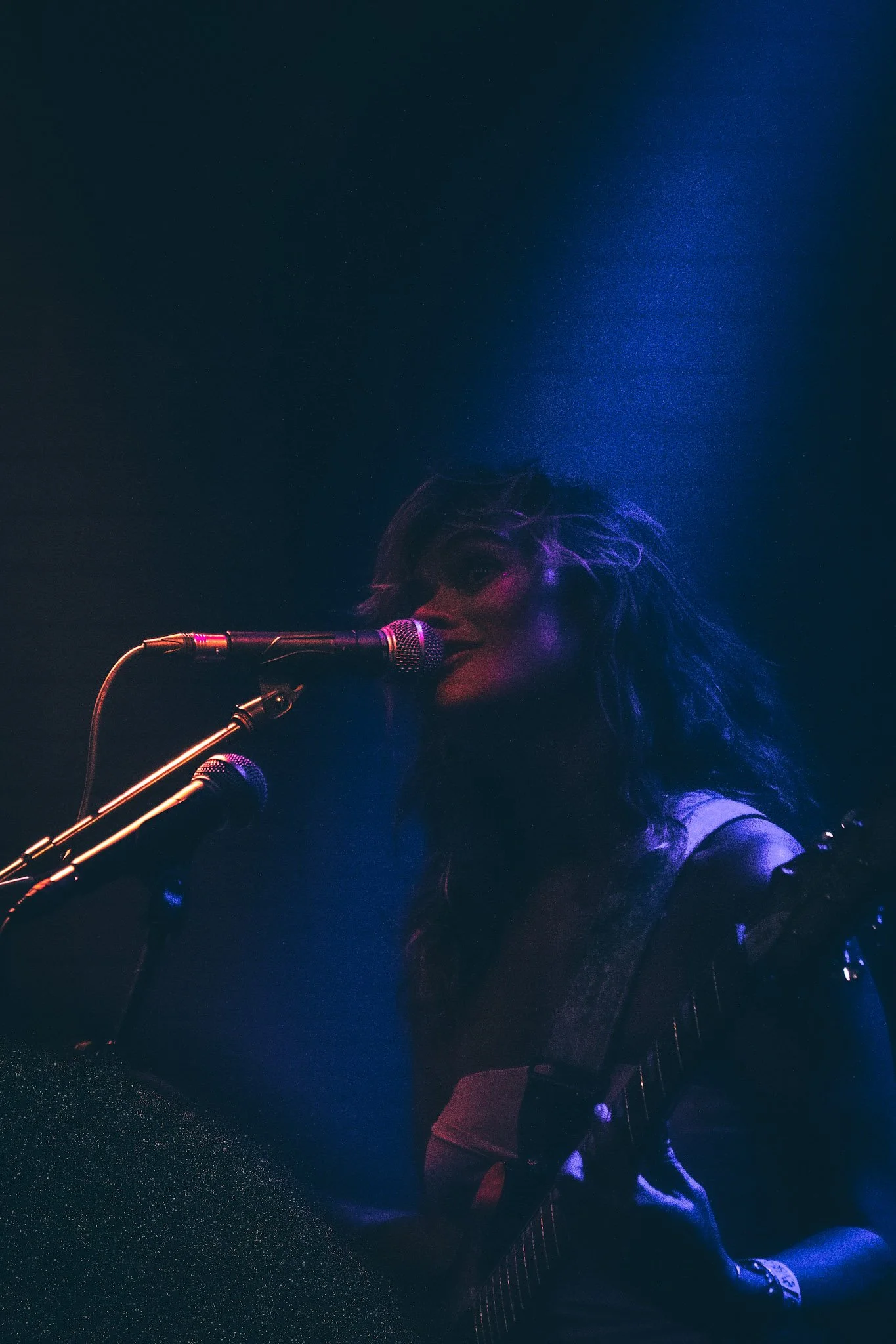 A musician sings into a microphone with colorful stage lighting, playing an electric guitar, with a dark background.
