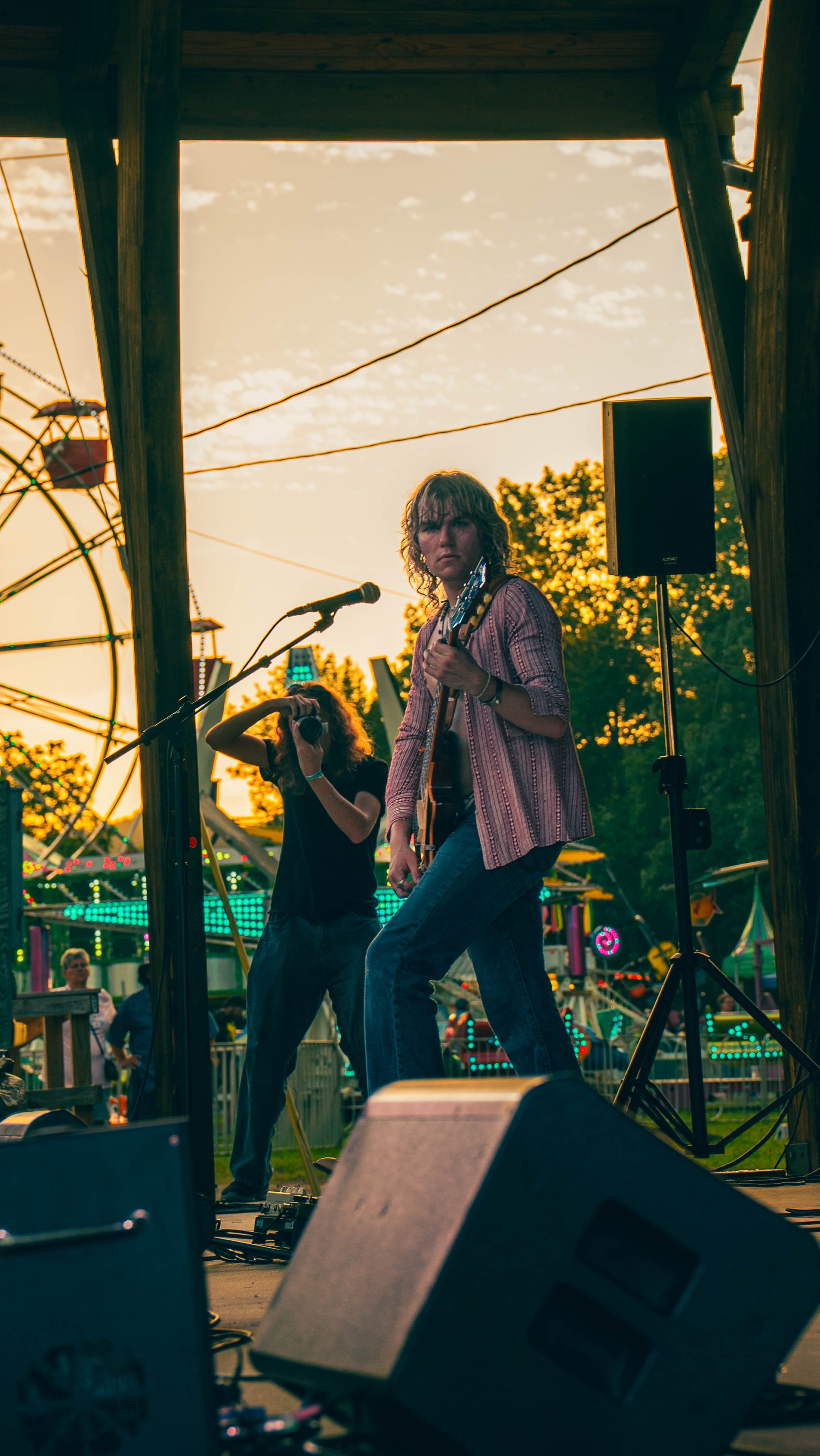 Young woman performing guitar on stage at outdoor carnival during sunset, with amusement rides and fairground lights in background.
