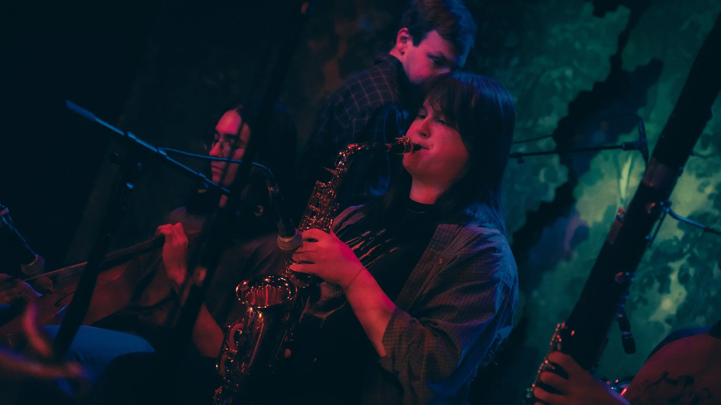 A group of musicians playing jazz instruments, with a focus on a woman playing the saxophone, in a dimly lit setting with colorful lighting and microphones.