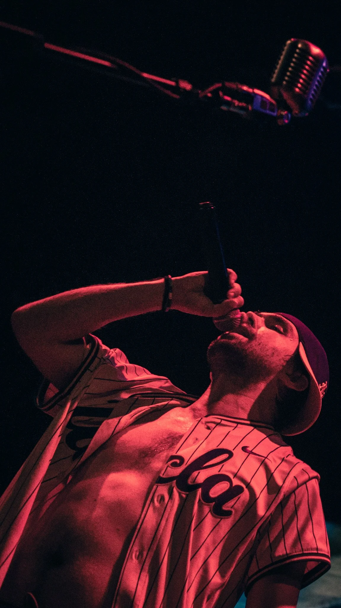 Male singer performing with microphone on stage at night, wearing baseball jersey and cap, illuminated by red stage lights.
