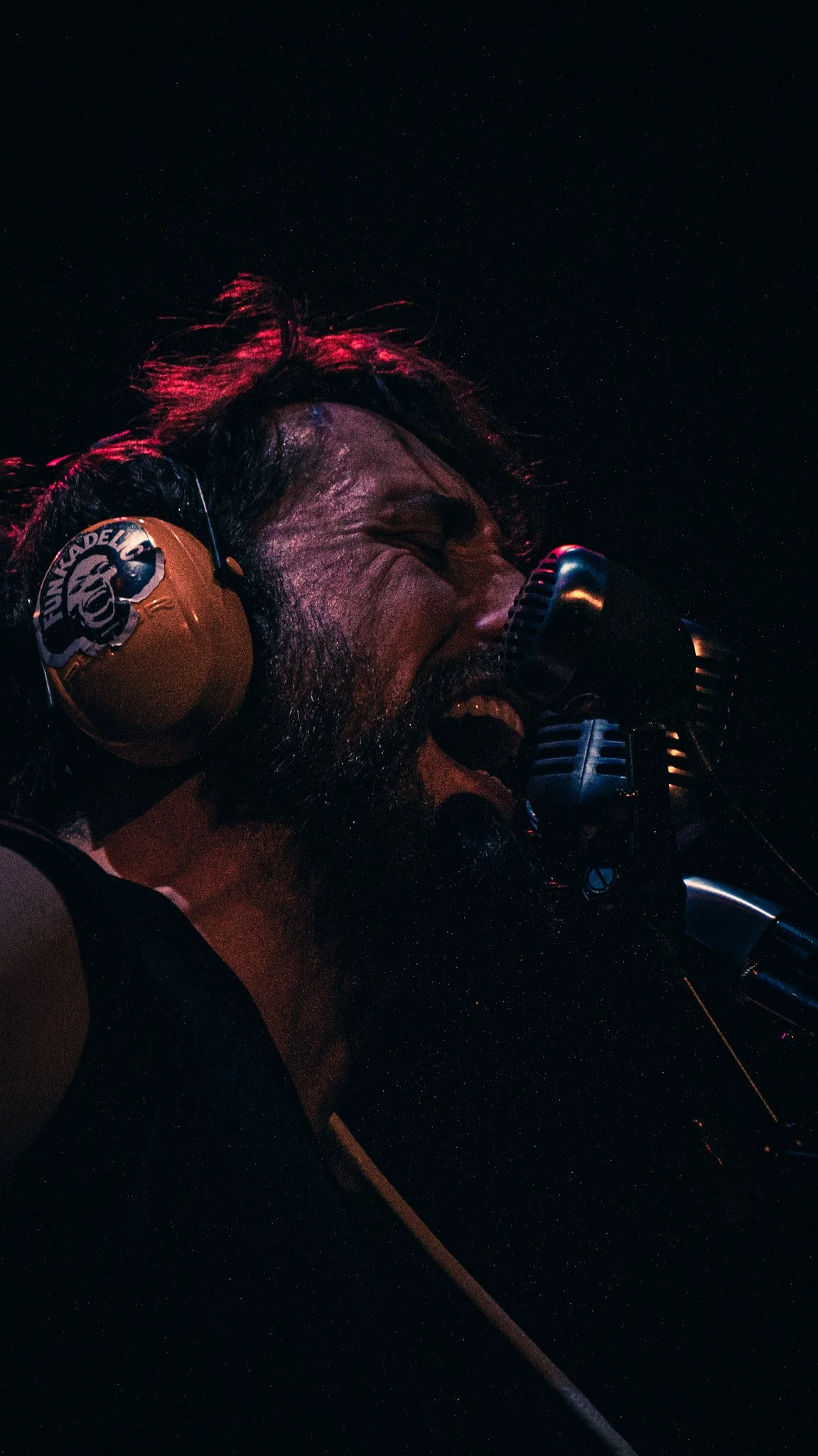 A man with long hair and a beard singing into a microphone, wearing headphones with the text 'FUNERAL' on them, in a dark environment with a starry background.