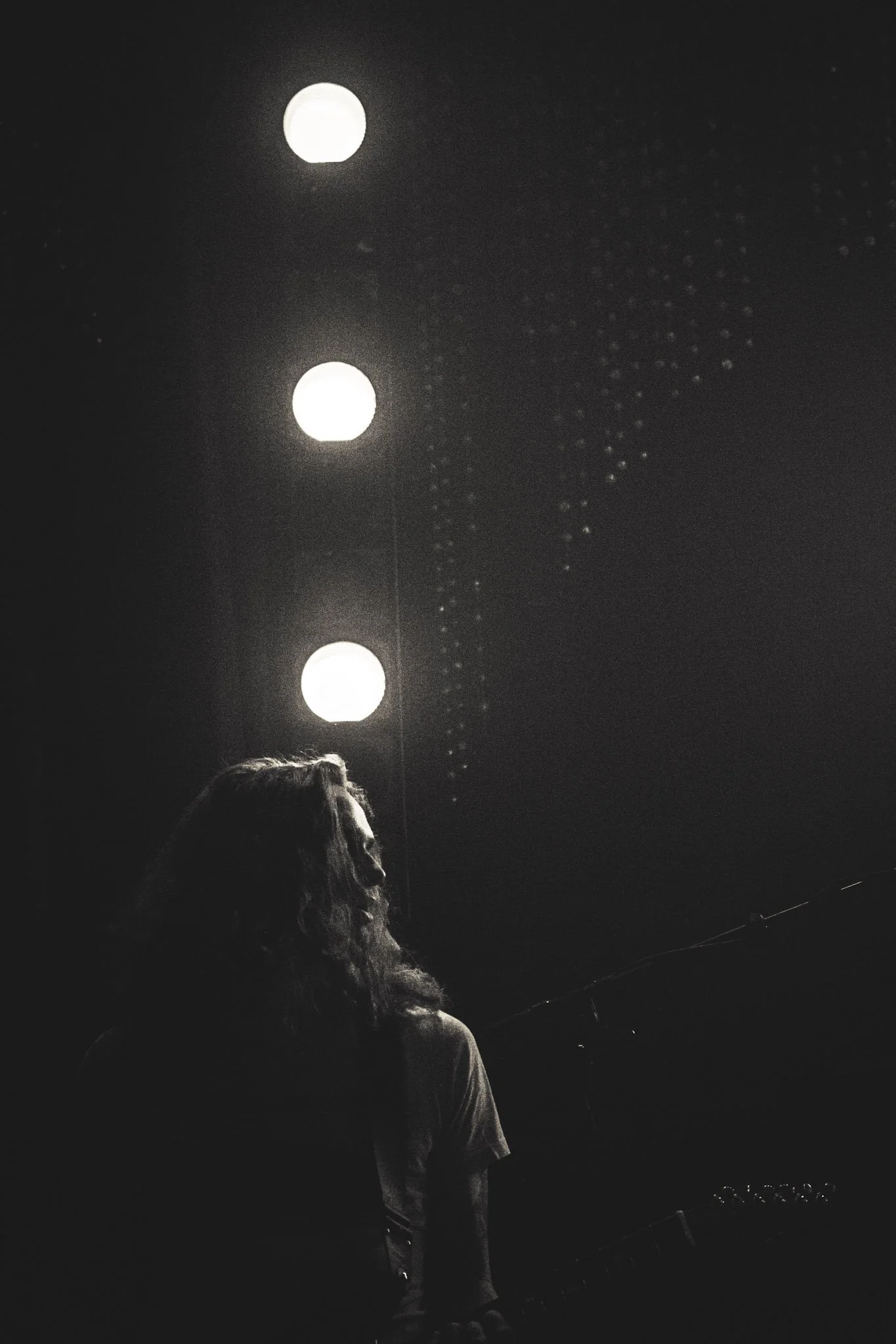 A woman with long hair stands in front of a dark background illuminated by bright stage lights. A row of small light bulbs is visible at the bottom right corner of the image.