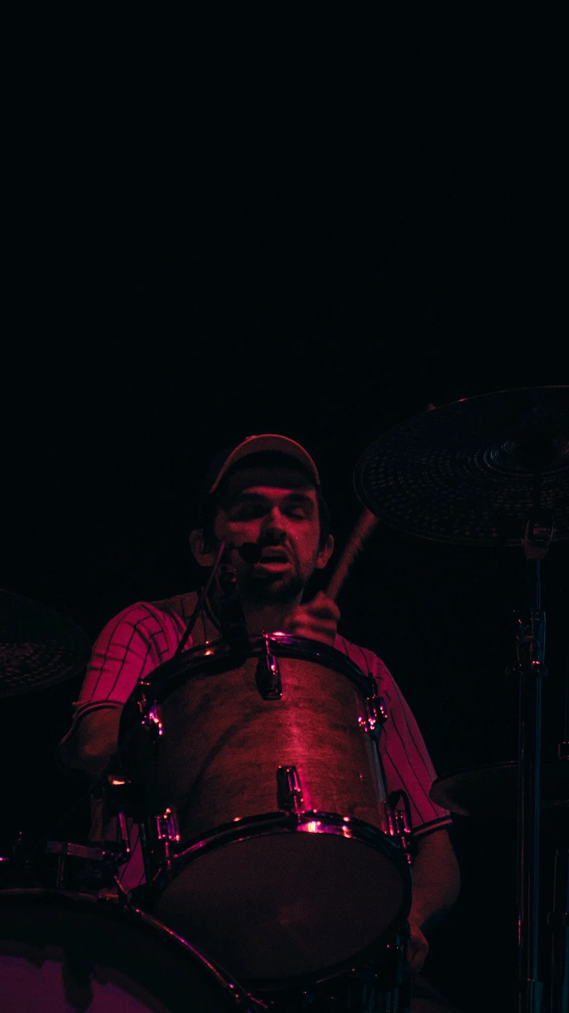 Man playing drums on stage with a dark background, illuminated by red lighting.