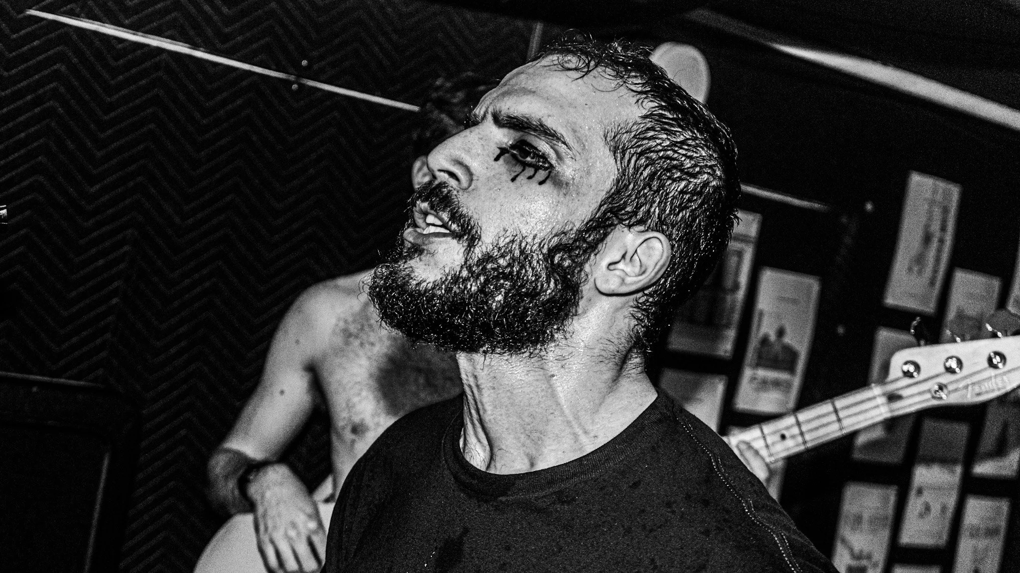 A black-and-white photo of a man with wet hair and a beard, singing or speaking passionately, with tattoos on his arm, in a music studio with acoustic foam walls.