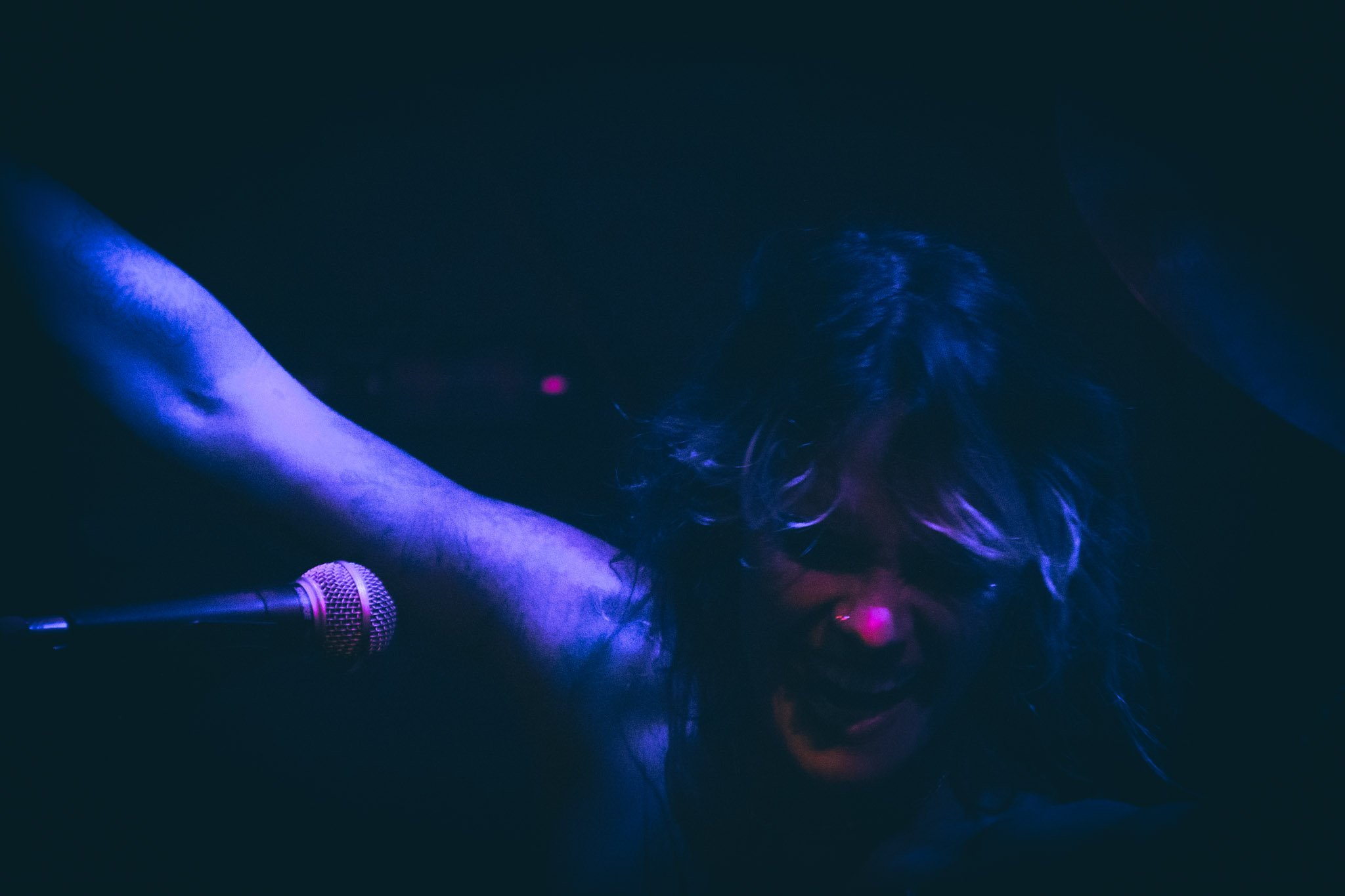 A performer with curly hair wearing makeup, in a dark setting, illuminated by blue and purple lights, holding a microphone.