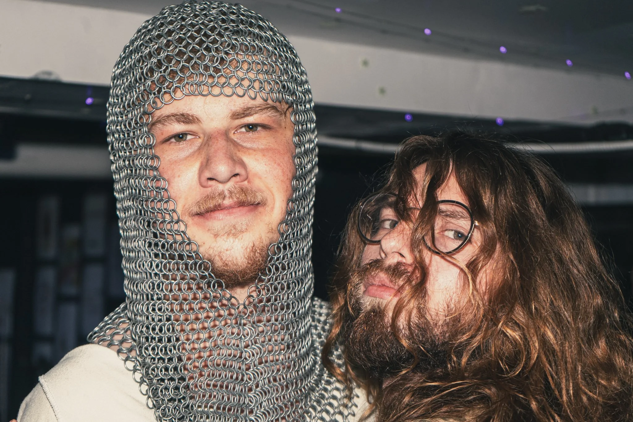 Two people close together, one wearing chainmail helmet with a serious facial expression and a man next to them with curly hair and glasses.