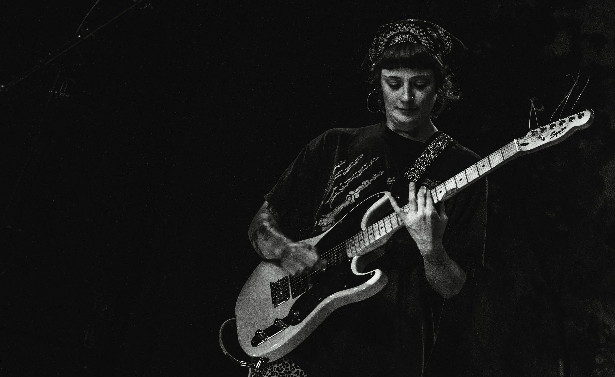 A woman playing an electric guitar on stage in a dark setting, wearing a bandana, large hoop earrings, and a black t-shirt.