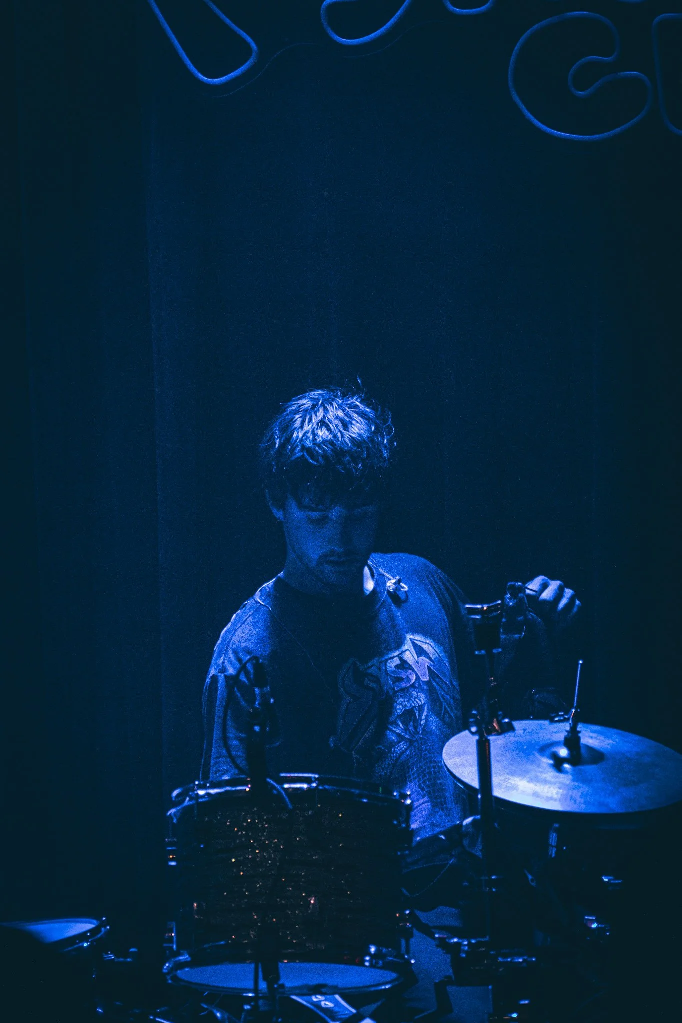 A person with curly hair playing a drum set in a dark room illuminated by blue light.