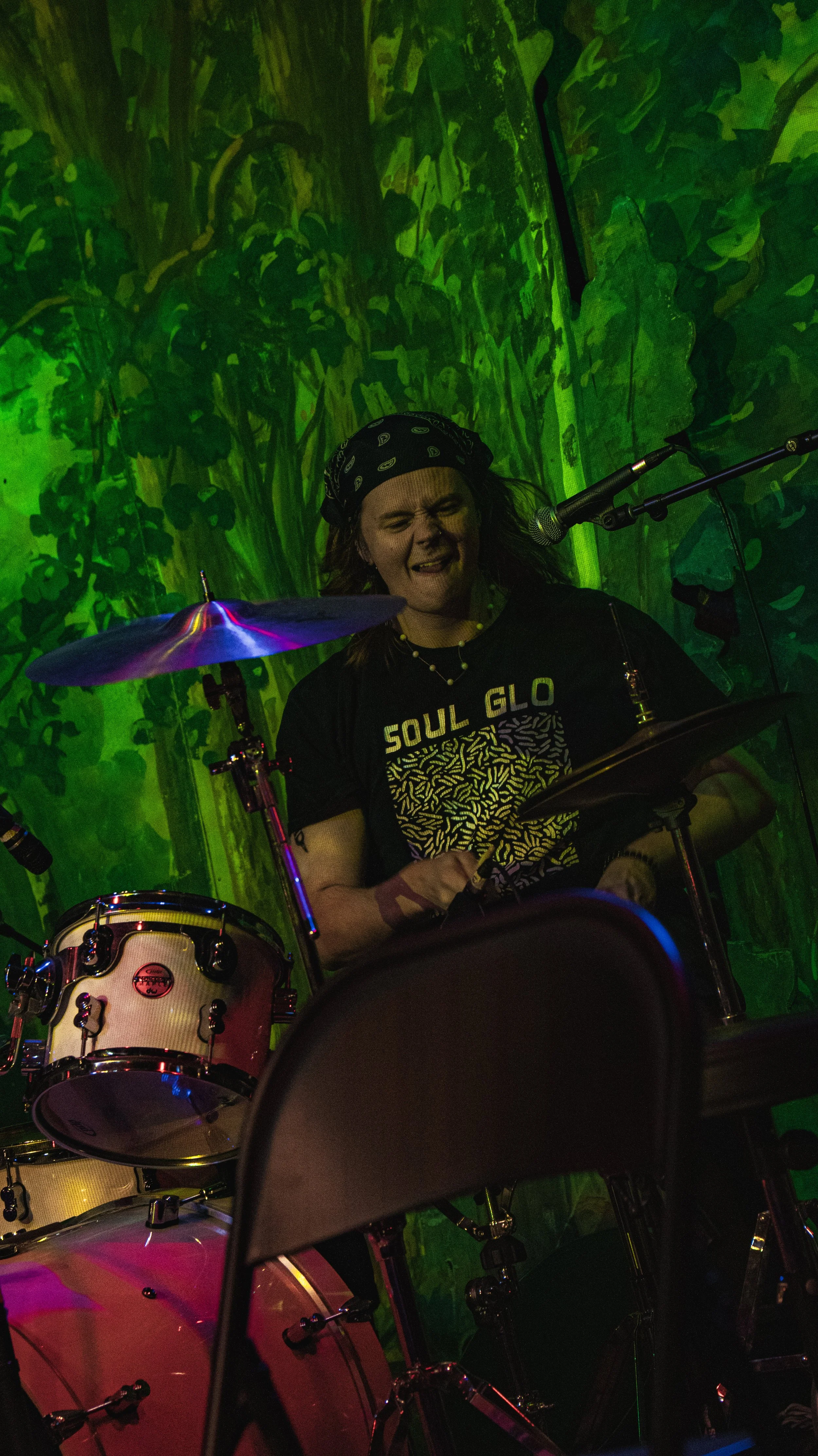 A woman with dark hair, wearing a black bandana and a black T-shirt with 'Soul Glo' printed on it, is playing drums in a dimly lit setting with green lighting and a painted backdrop of trees.