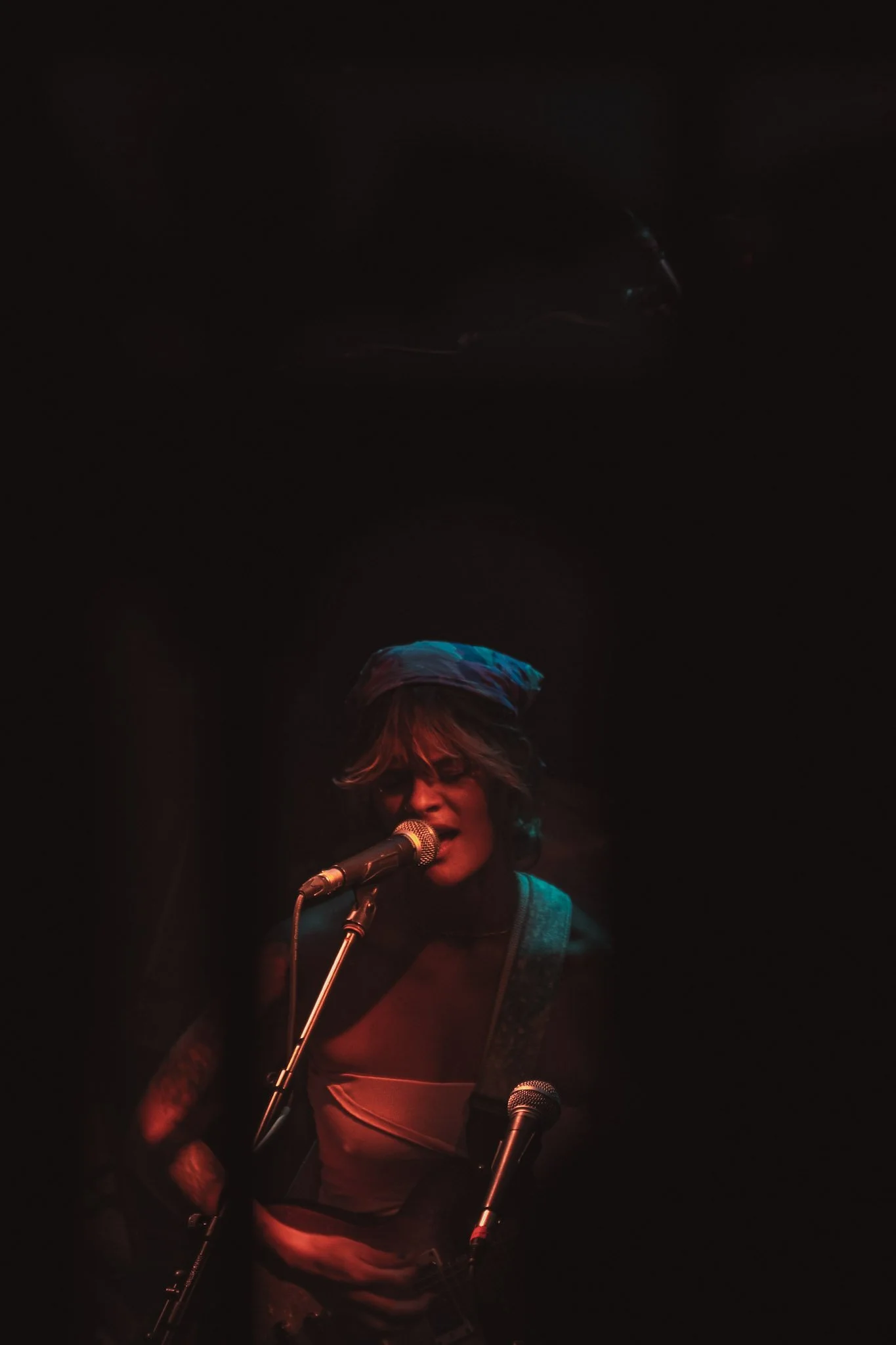 A woman with short hair and glasses singing into a microphone on a dimly lit stage.