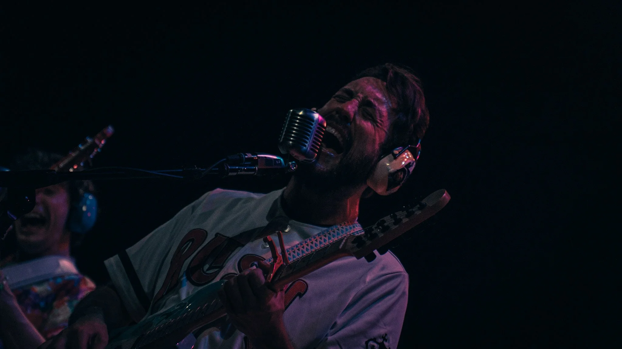 A man wearing headphones and a white sports jersey singing or speaking into a vintage microphone, with another person partially visible, holding a guitar in a dark setting with colorful lighting.