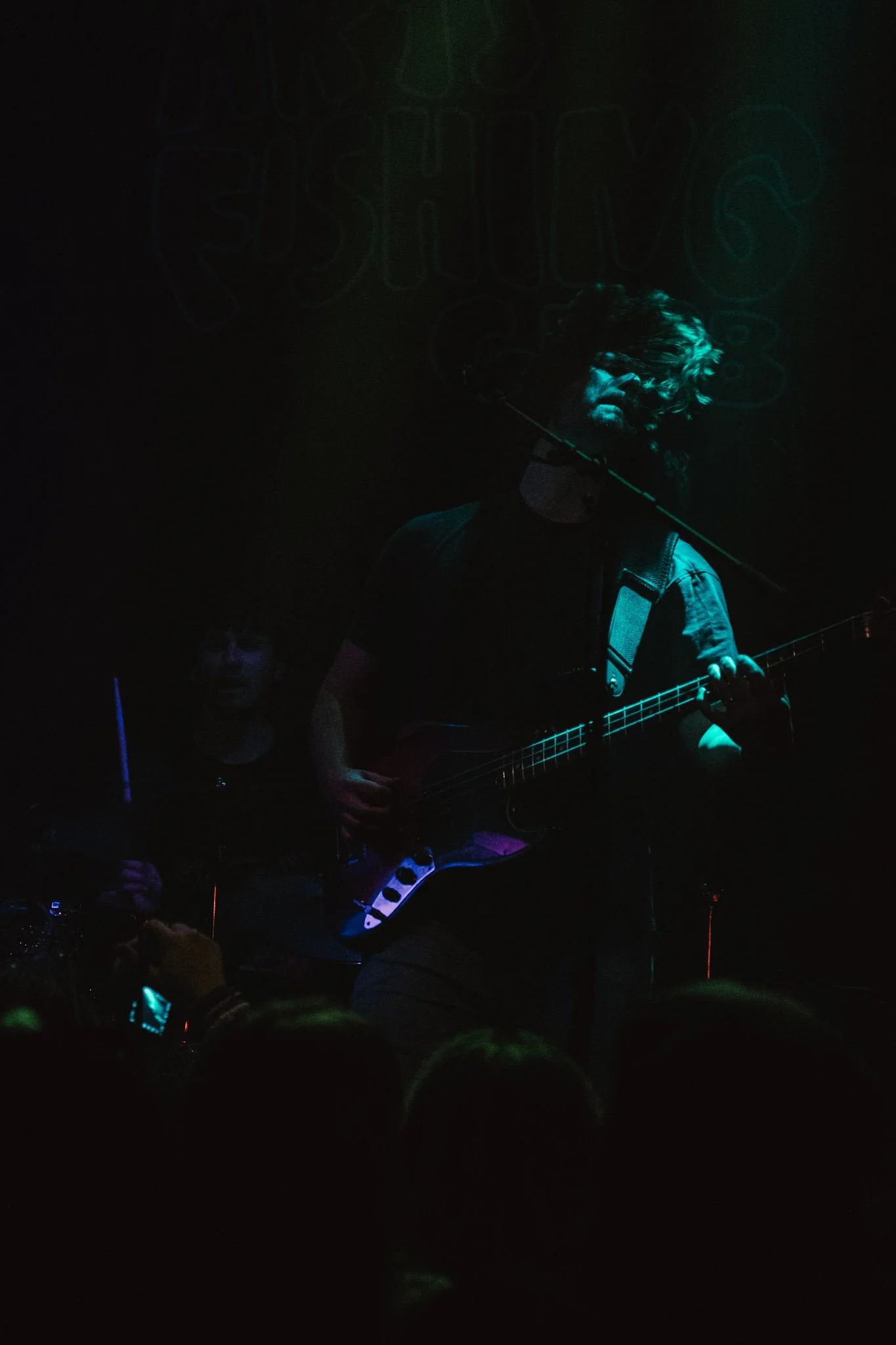 A person with long curly hair singing into a microphone and playing a guitar on stage with dark lighting and a illuminated background.