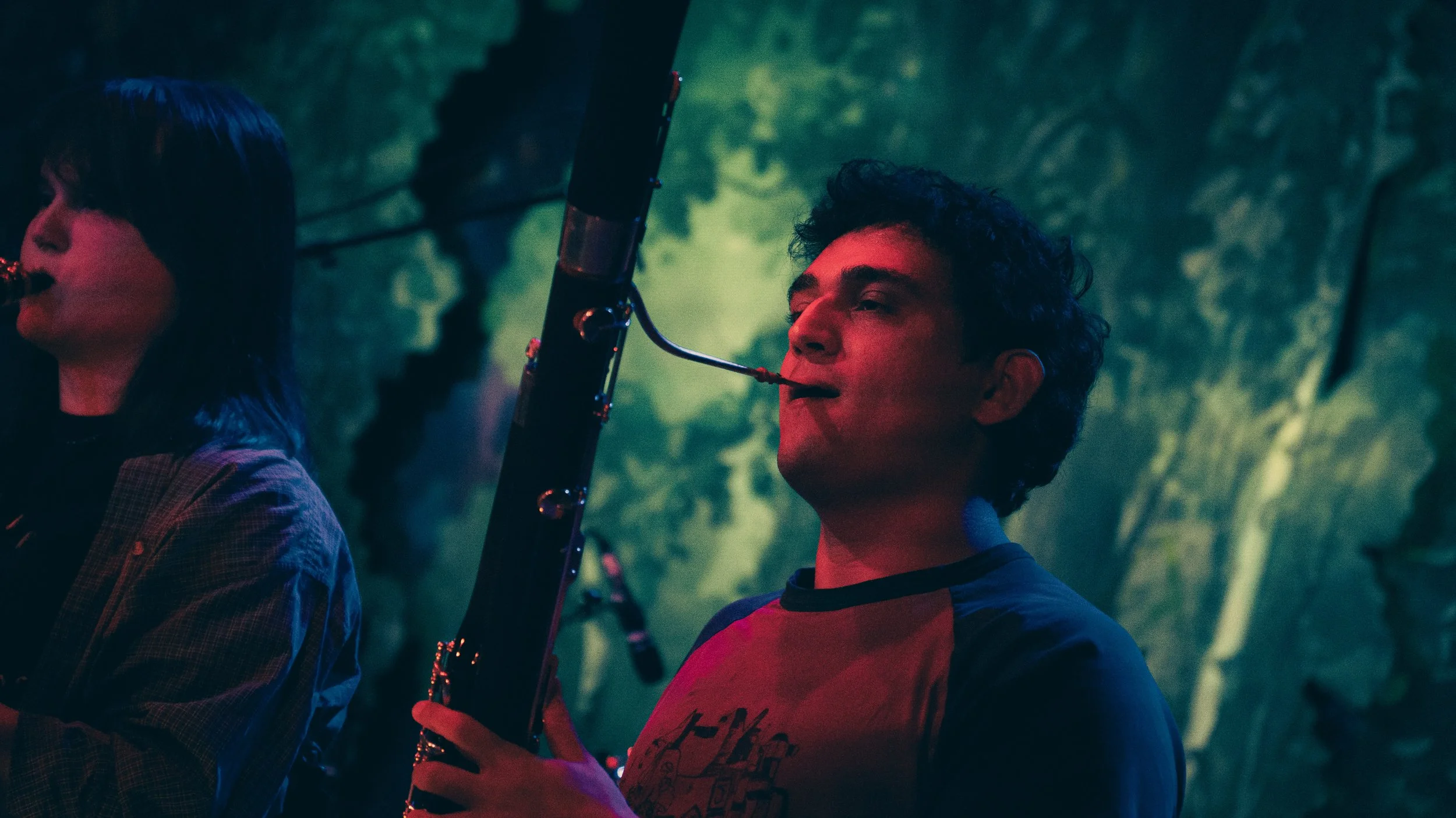 A young man with curly hair playing an electric guitar in a dimly lit, outdoor setting with green and blue lighting, alongside a woman playing a bass guitar, both immersed in music.