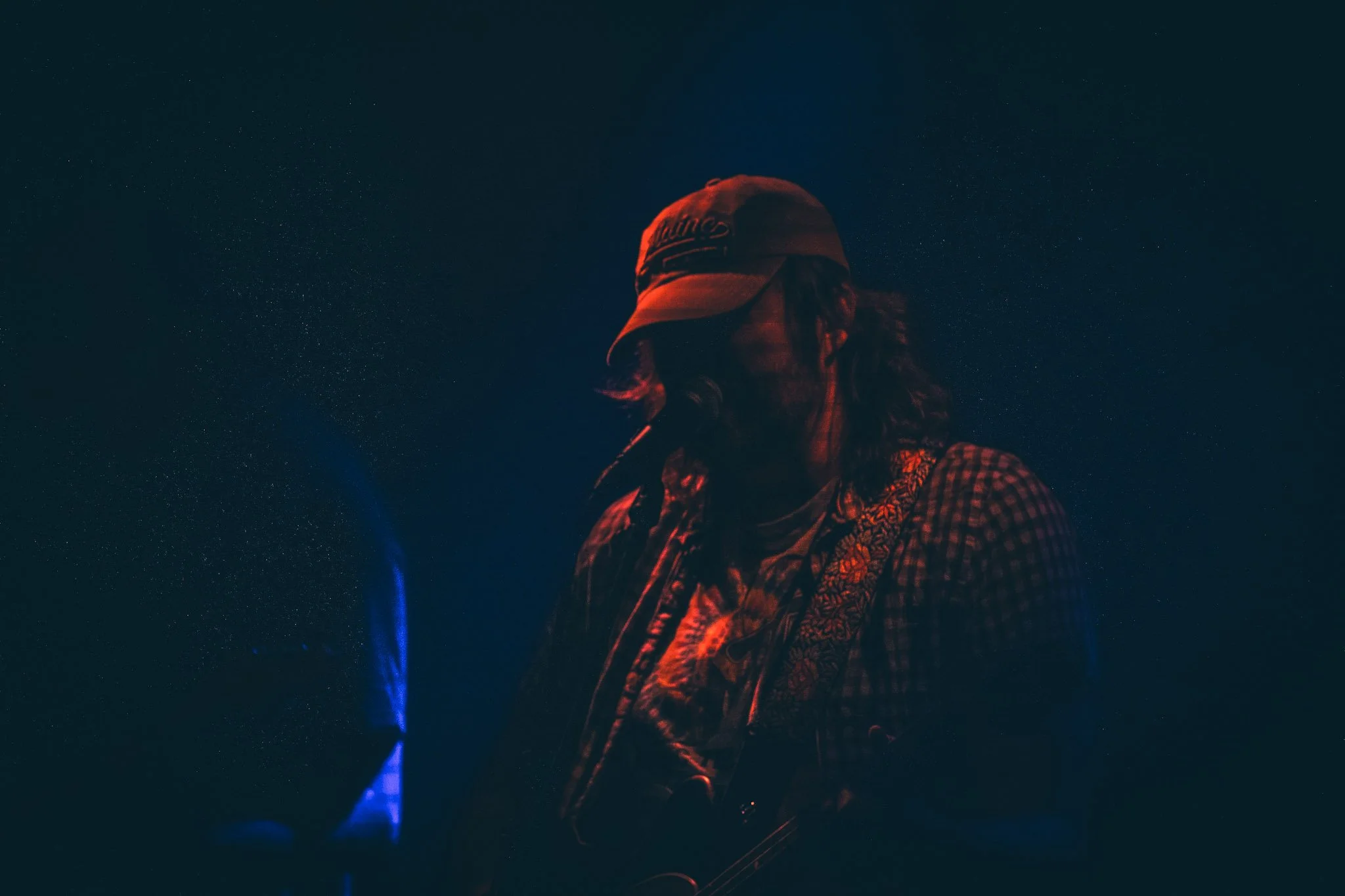 A person with long hair wearing a baseball cap and a plaid shirt, seen against a starry night sky, illuminated by red and blue lighting.