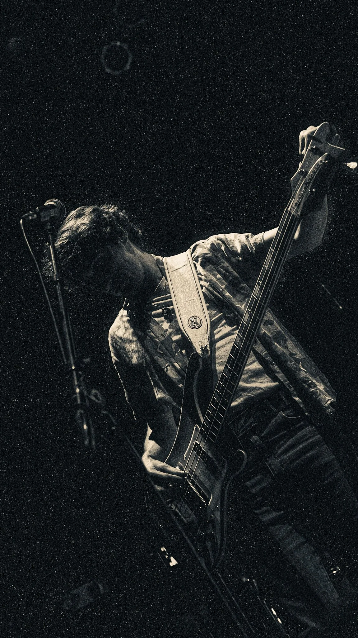 A person playing an electric bass guitar on stage in black and white. The individual is wearing a plaid shirt and glasses, and is connected to a microphone stand.