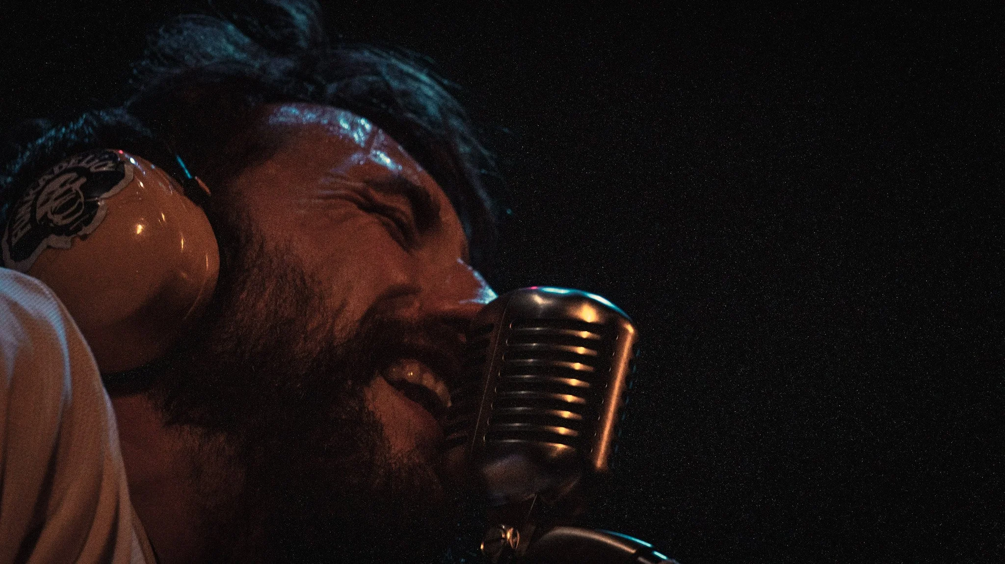 A man with dark hair and a beard singing into a vintage-style microphone against a dark background.