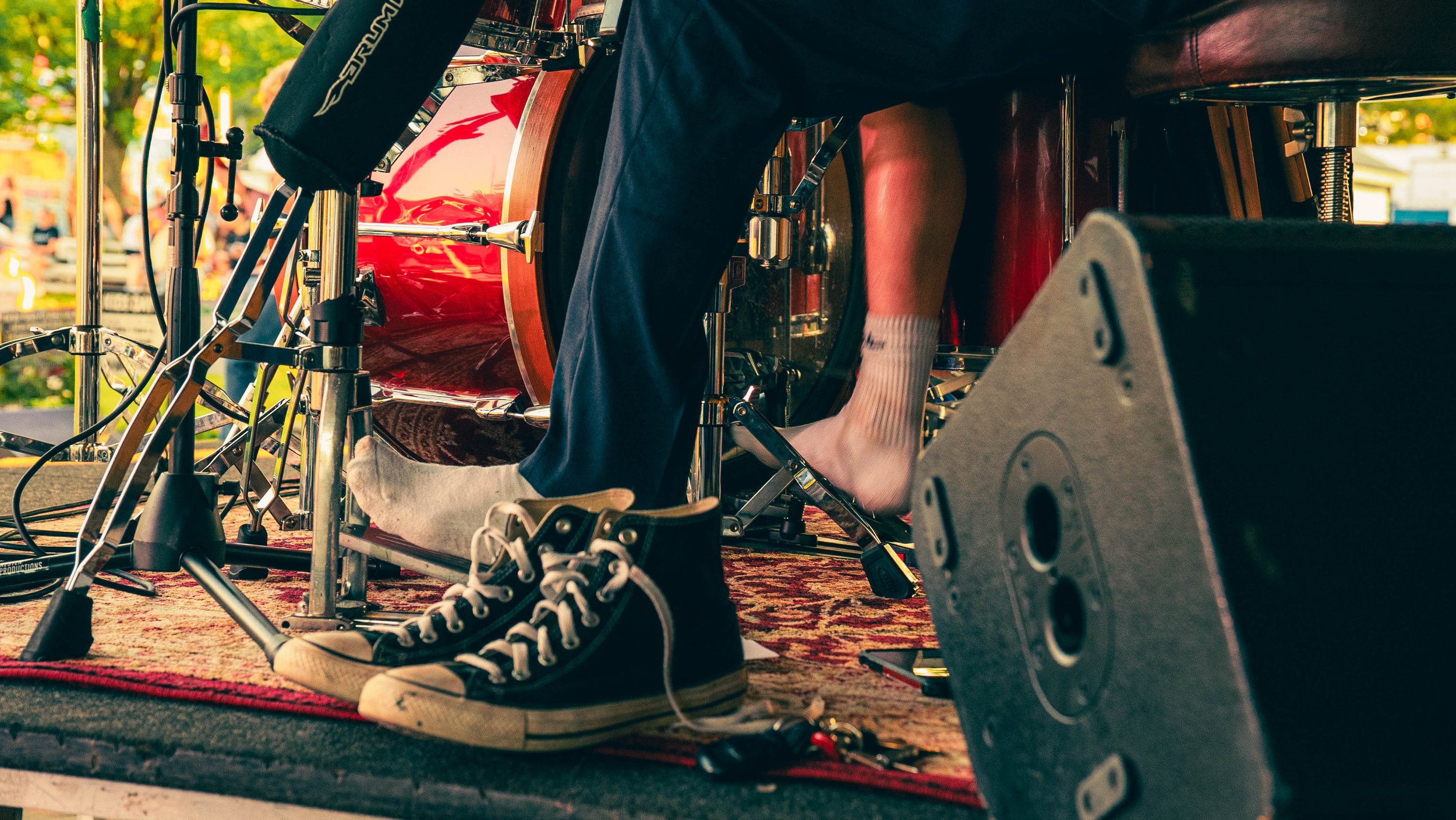 A musician playing drums on an outdoor stage, visible from low angle, with focus on their legs, drum set, and sneakers.