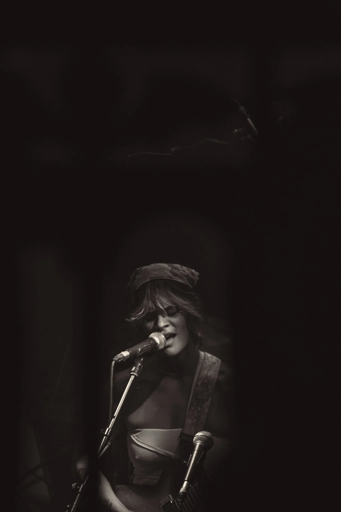 A person with short hair and glasses is singing into a microphone and playing a guitar on stage, illuminated by stage lighting in a dark setting.