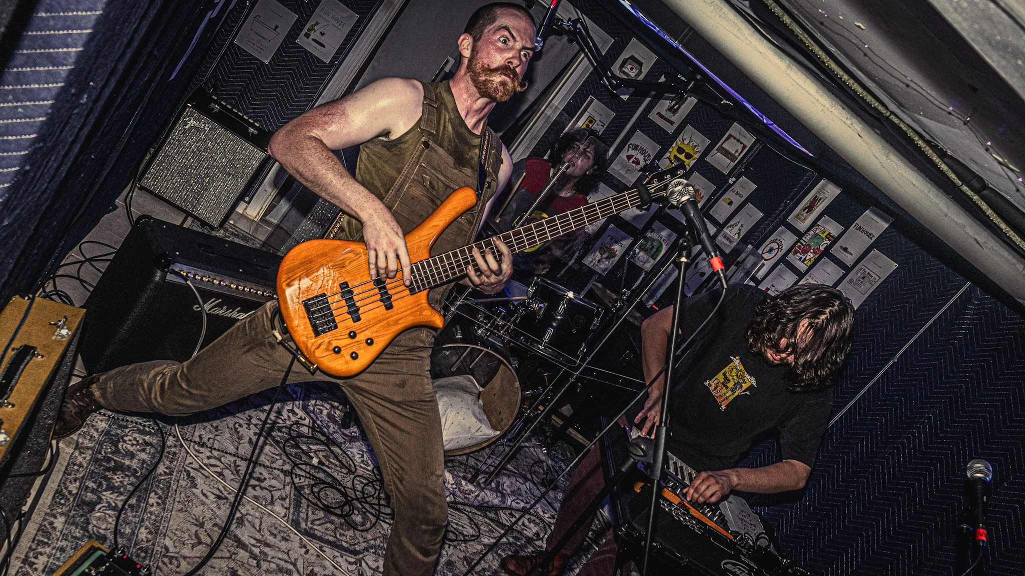 A musician with a beard and mustache, wearing a sleeveless shirt and brown pants, playing an orange electric bass guitar on stage. Behind him, another band member is playing a keyboard with long hair, in a room decorated with posters and music equipm