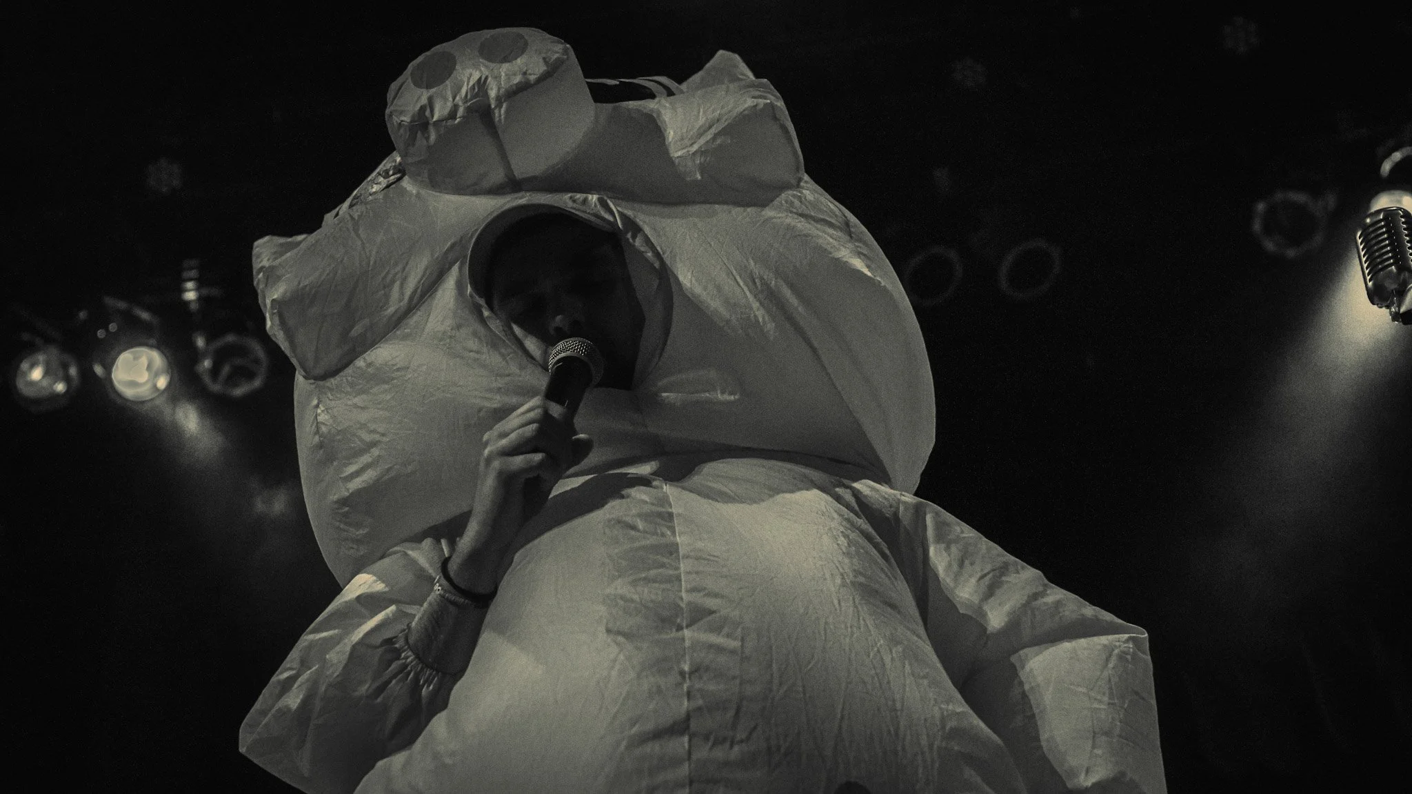 Performer in a white protective suit holding a microphone on stage, with stage lights in the background.
