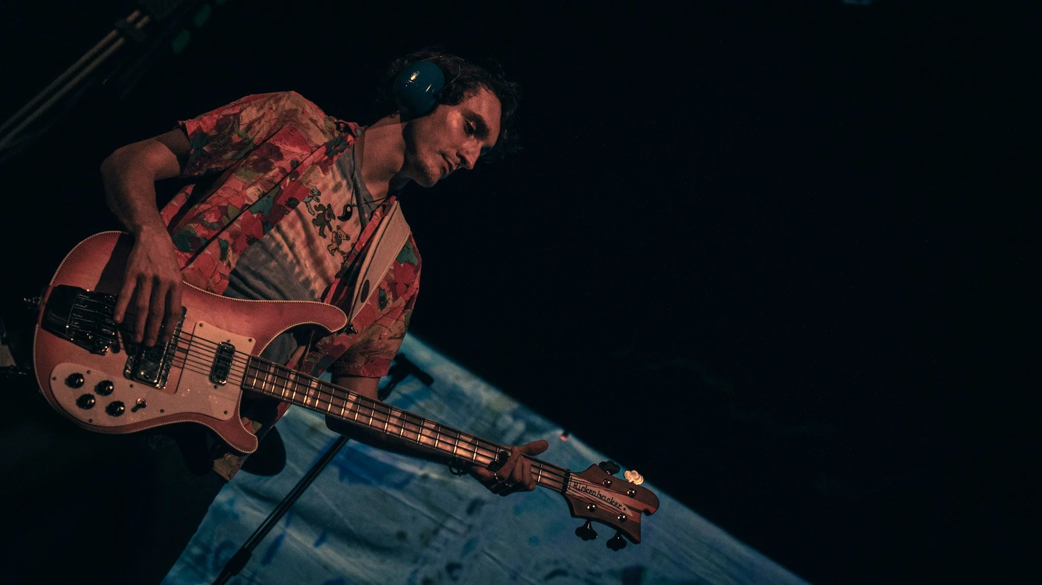 A man wearing a colorful short-sleeved shirt with a floral pattern, headphones, and holding a pink electric guitar, performing on stage in a dimly lit setting.