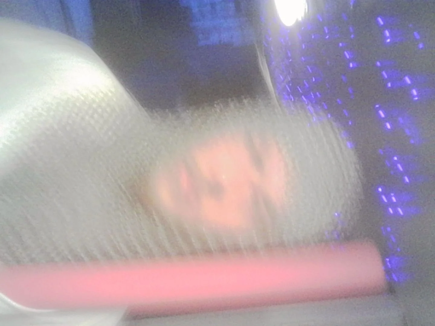 Close-up view through a textured surface or screen showing a blurred reflection of a face with a pinkish hue, with blue and purple lights in the background.