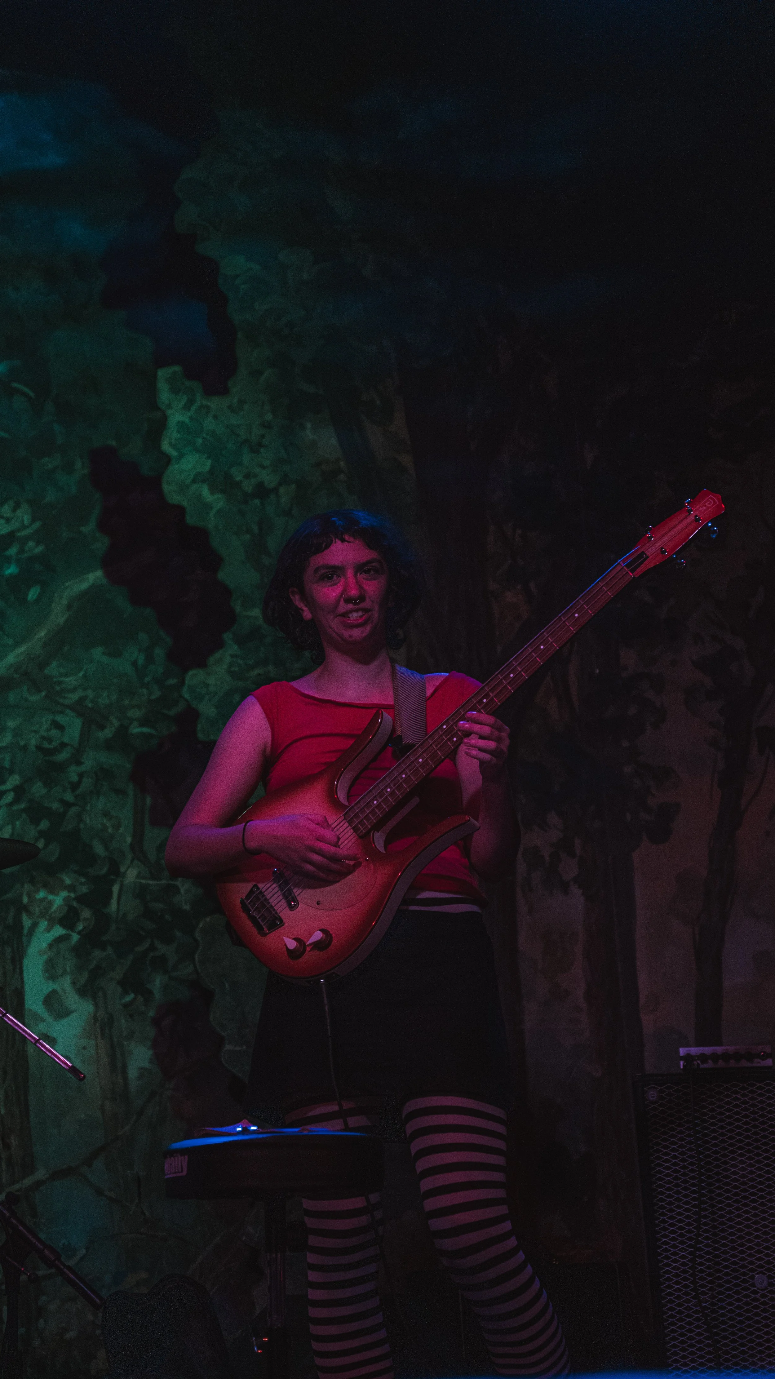 A woman playing an electric guitar on stage with a backdrop that looks like a forest. She has short dark hair, is wearing a red sleeveless top, a black skirt, and black-and-white striped tights. She appears to be smiling and engaged in her performanc