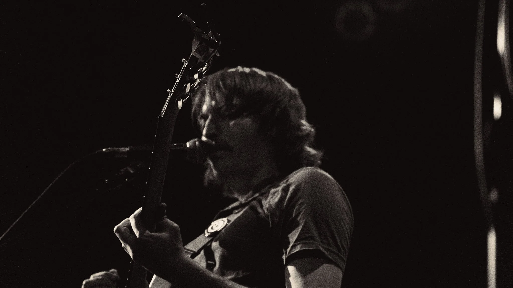 A person with long hair, wearing a jacket with a patch, is playing the guitar on stage in a dark setting.