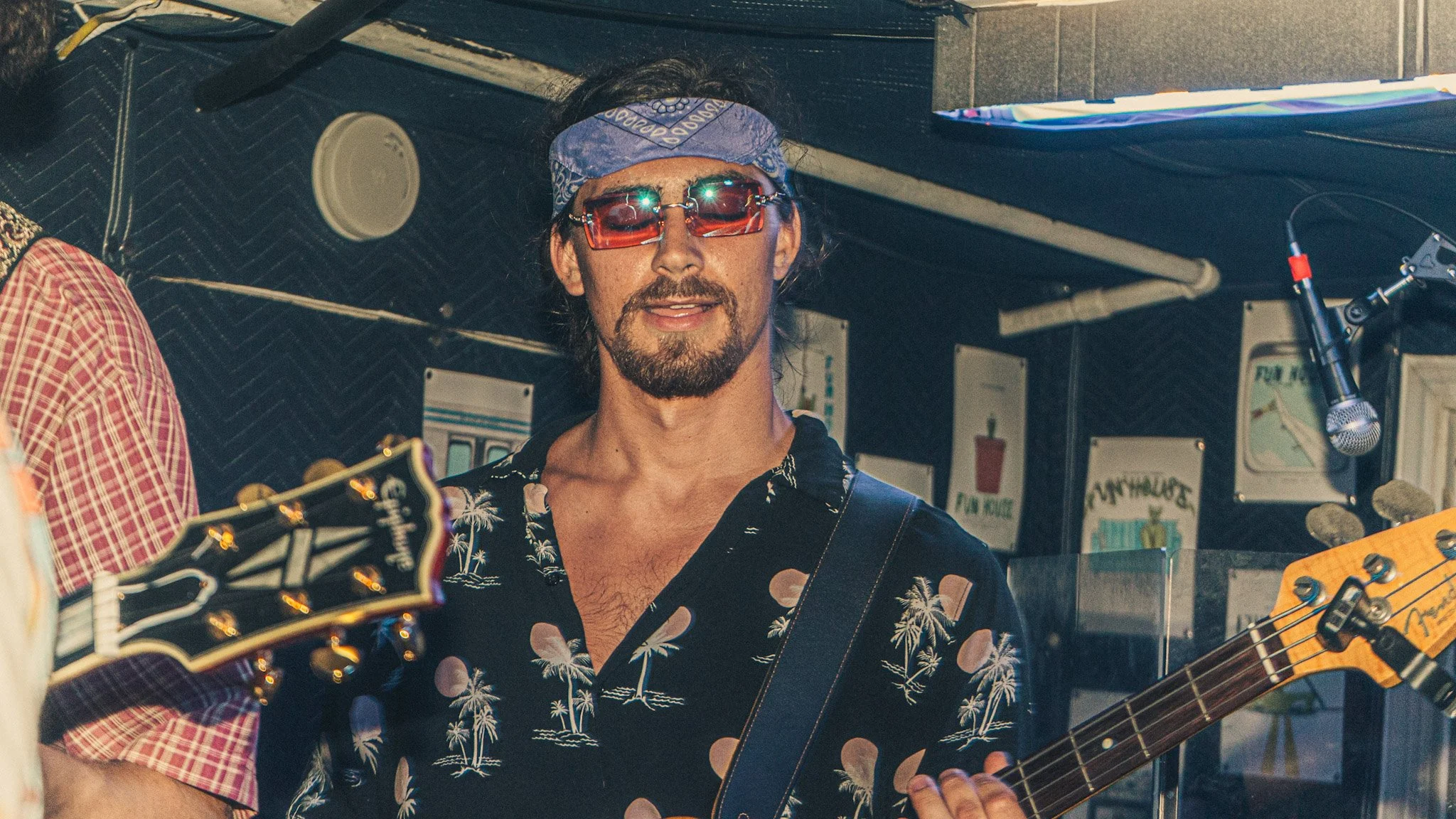 A man wearing red sunglasses and a blue bandana playing an electric guitar at a music venue. He has a beard and mustache, and a black Hawaiian shirt with palm tree patterns.