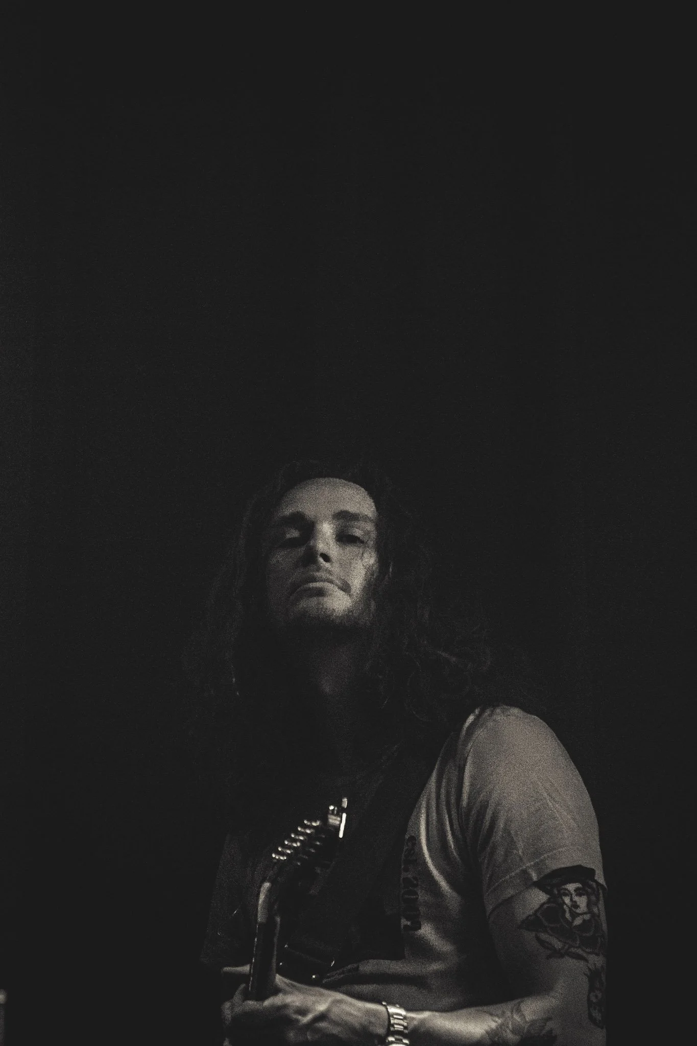 A woman with dark curly hair, tattoos on her arm, and wearing a watch, is taking a selfie in a dark setting with only her facial features and partial upper body visible.