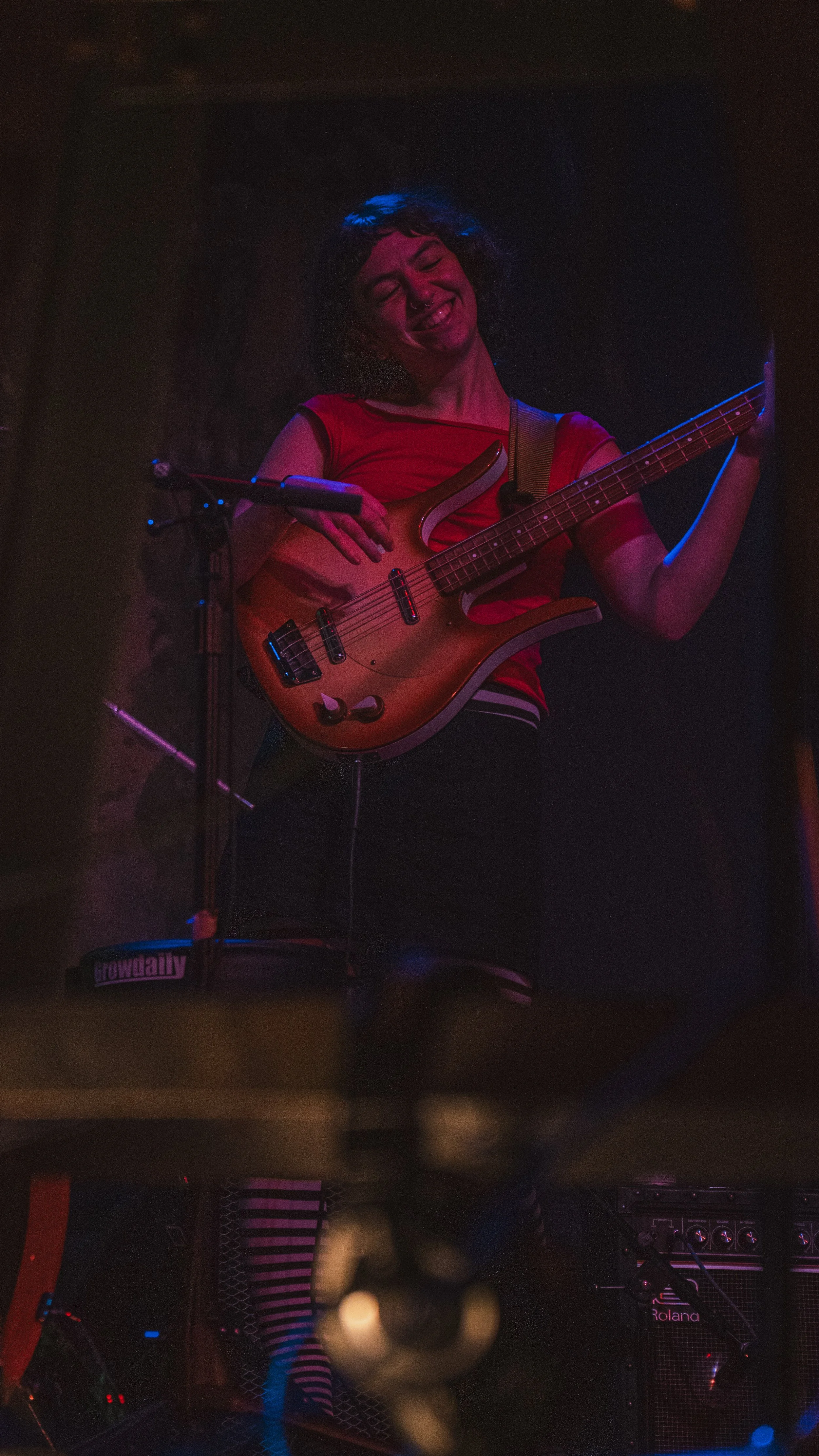 A person with curly hair playing an electric bass guitar on stage, smiling, with dark background and stage lights.